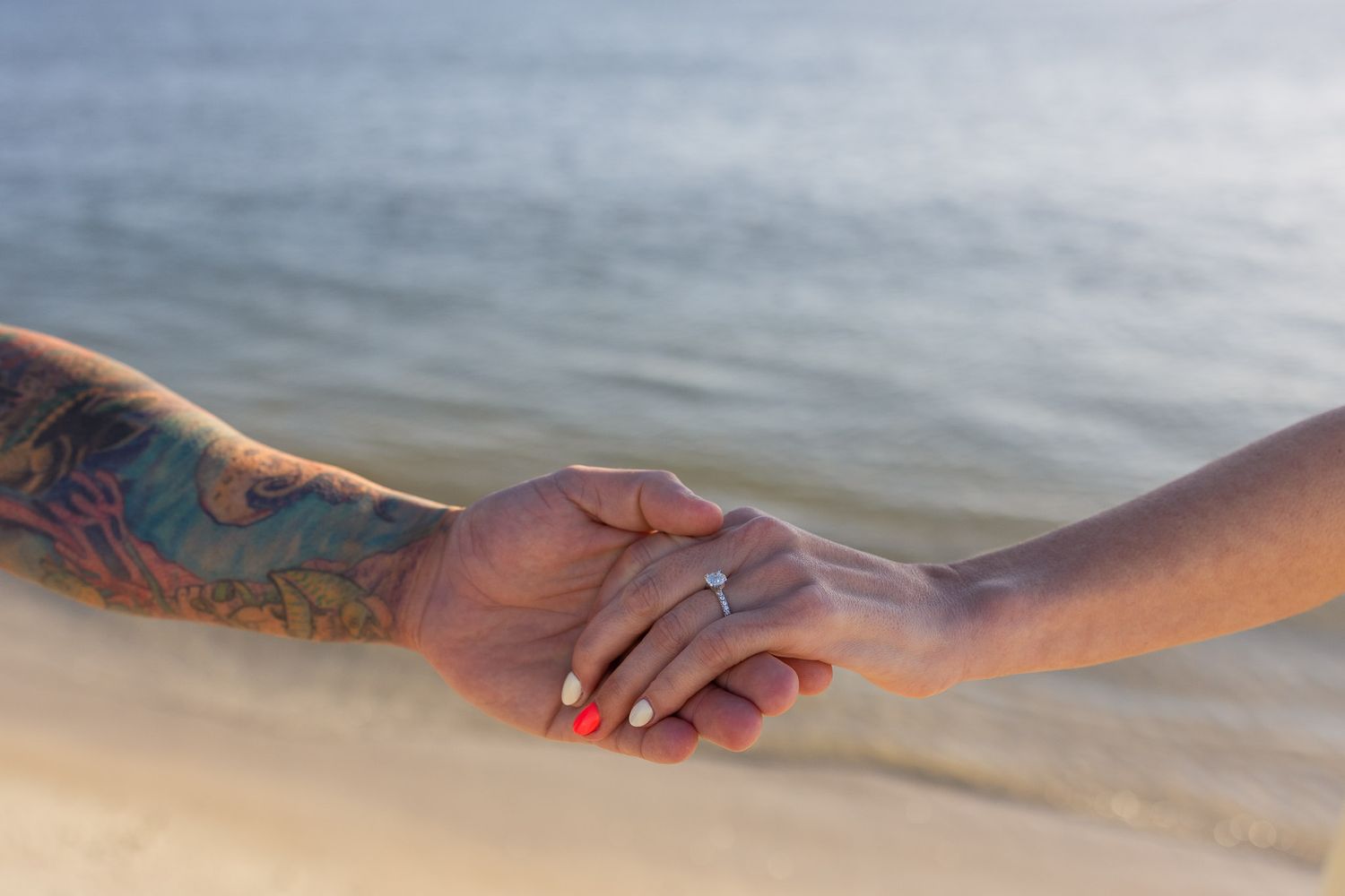 Two hands holding at the beach, one with colorful tattoos and the other wearing a diamond engagement ring.