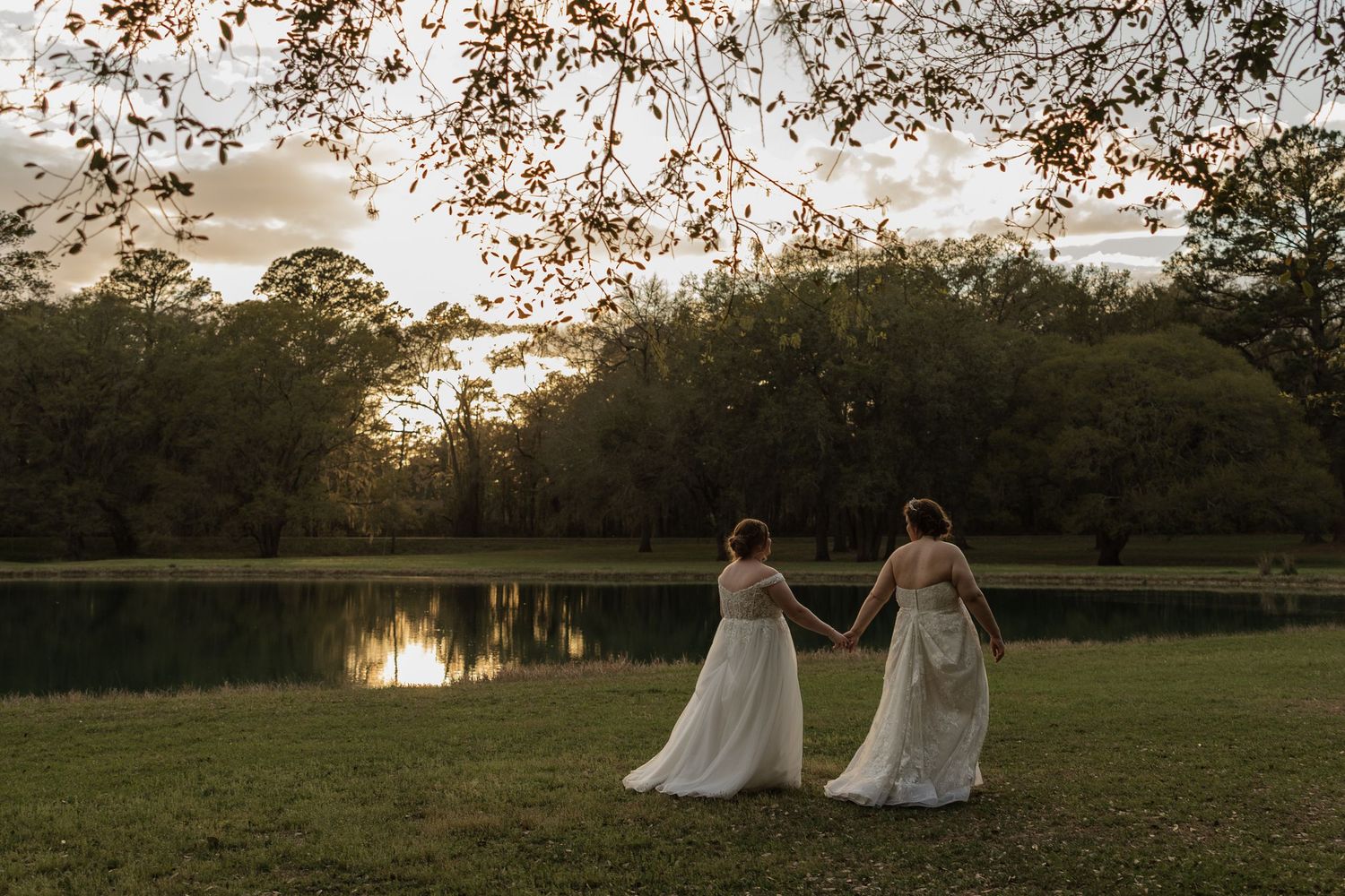 Two brides in white gowns hold hands by a serene lake at sunset surrounded by lush green trees.