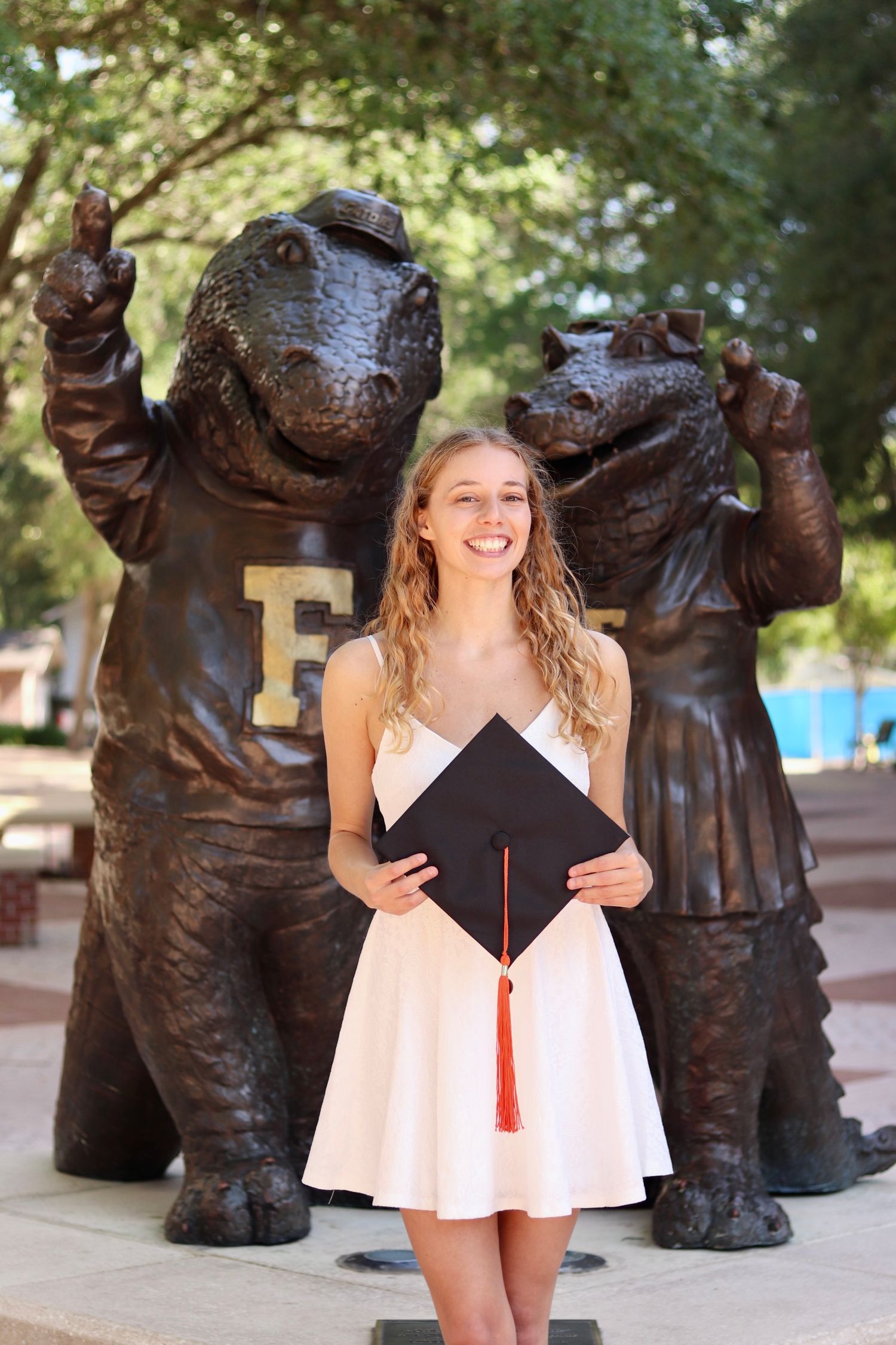 Graduate poses with bronze bear statues in a white dress while holding a graduation cap with red tassel.