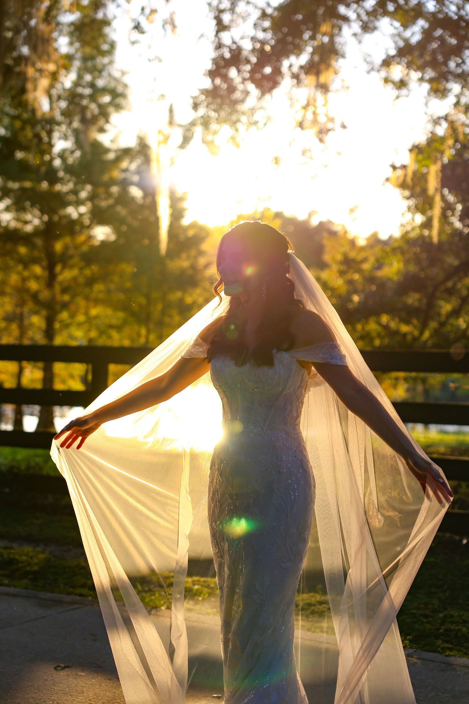 Bride in white dress poses with flowing veil backlit by golden sunlight in outdoor setting.