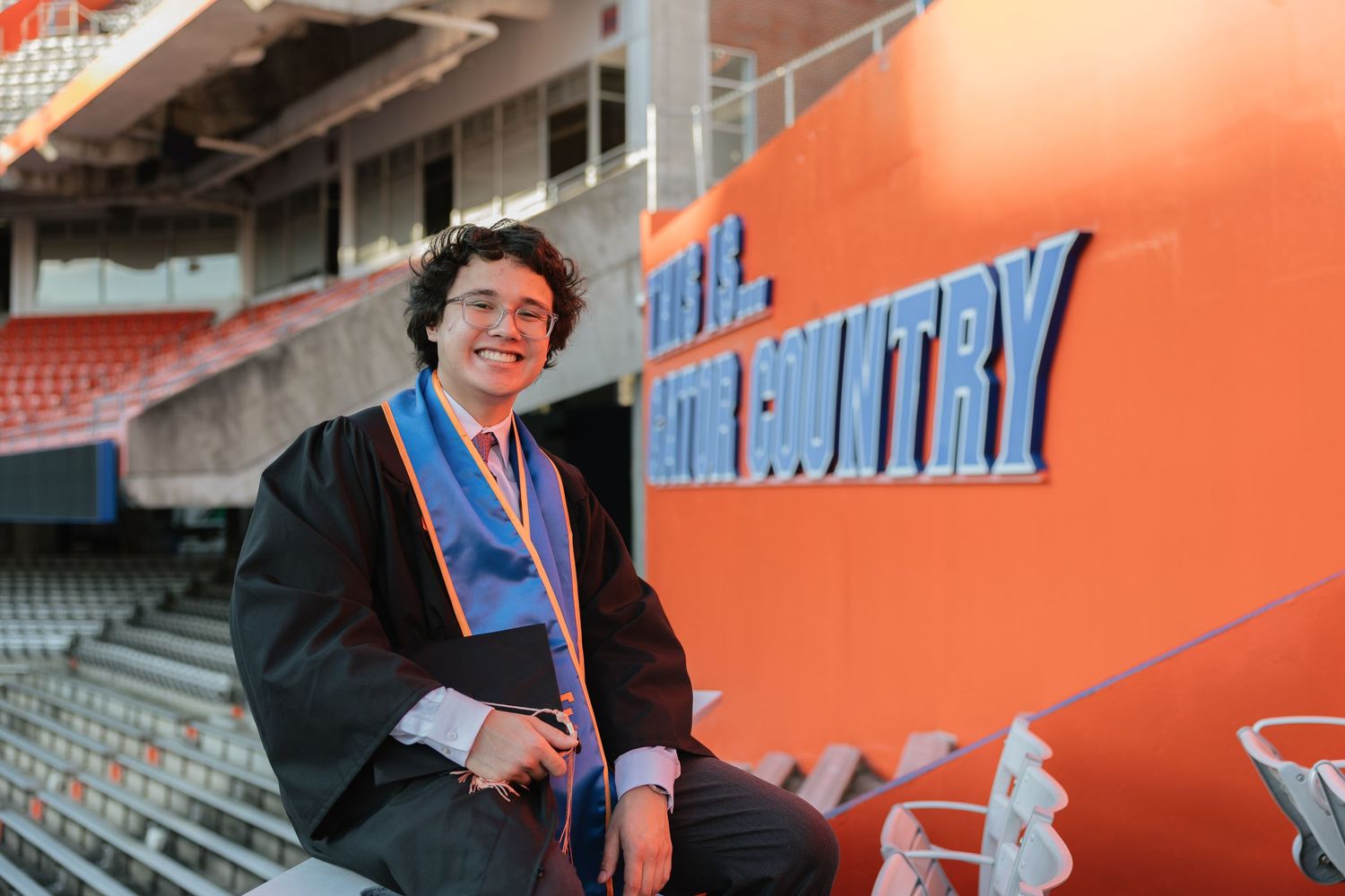 Graduate in cap and gown smiles in stadium seating beside orange Gator Country wall at University of Florida.