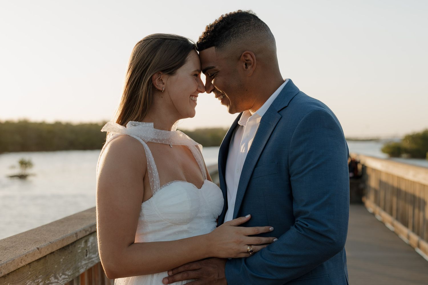 Couple in wedding attire sharing romantic moment on waterfront bridge at sunset, groom in blue suit, bride in white dress.