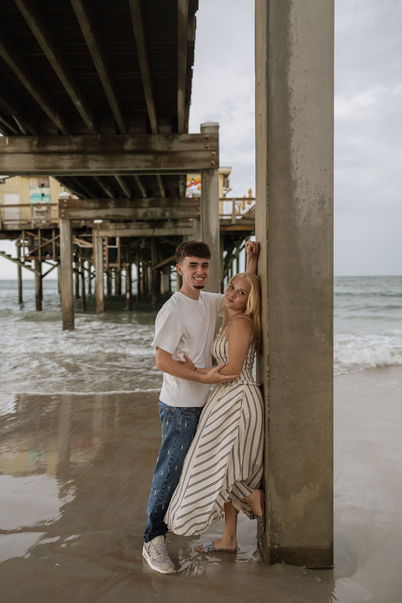 Couple embracing under a beach pier, woman in striped dress leaning on pillar, waves in background.