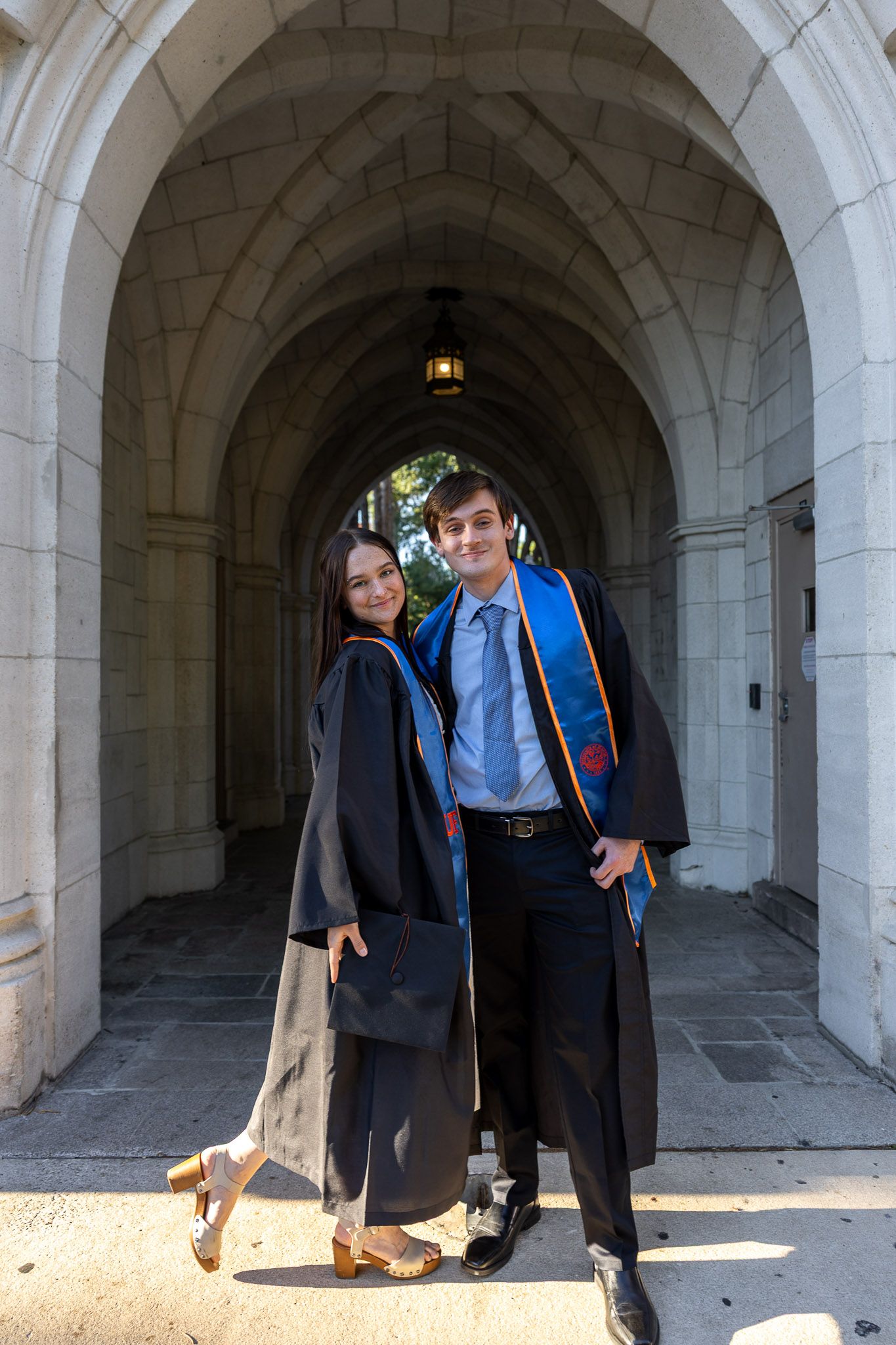 Two graduates in black academic robes pose under a stone archway, smiling for a graduation photo.
