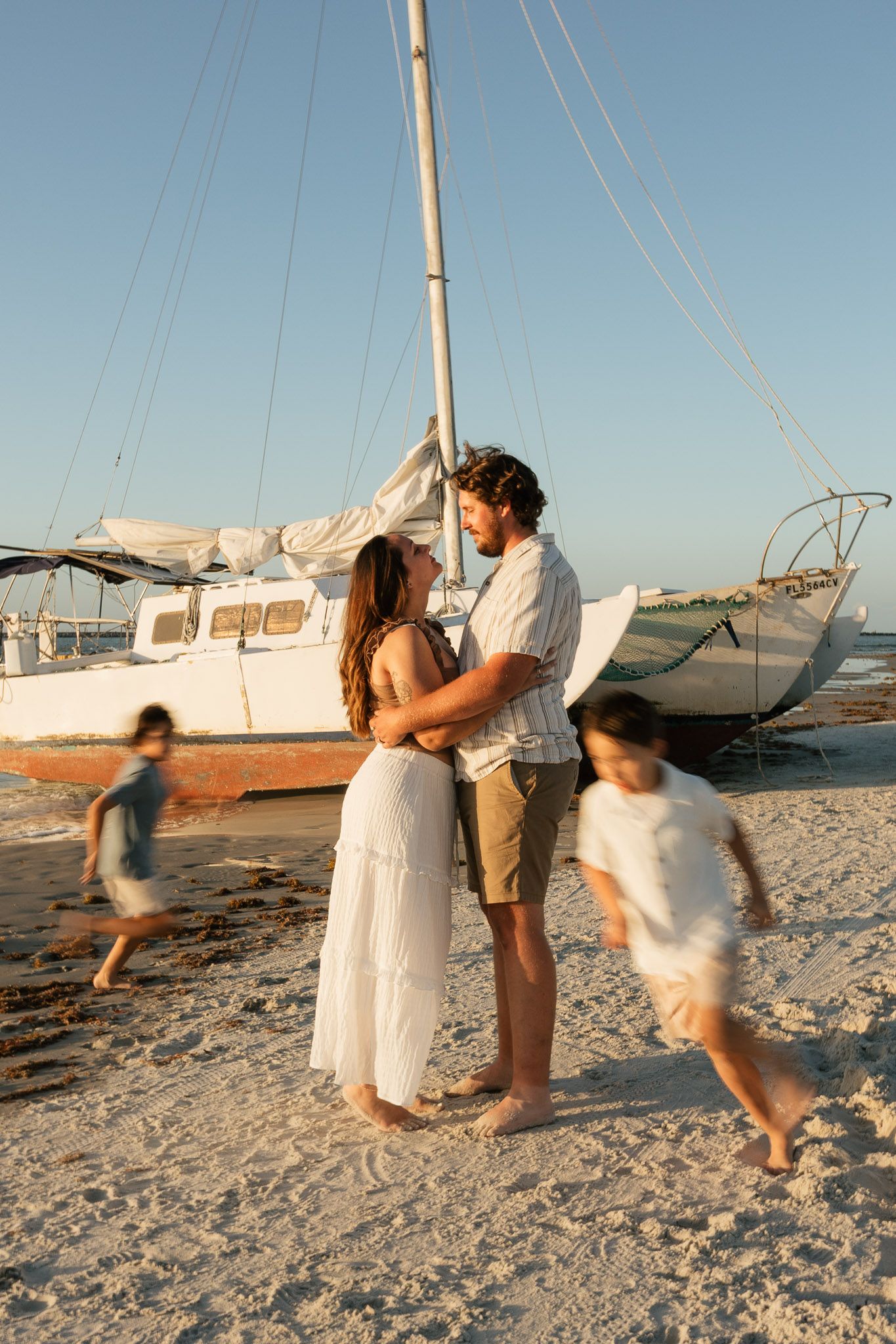 Couple embracing on sandy beach near sailboat at sunset while two children play around them.