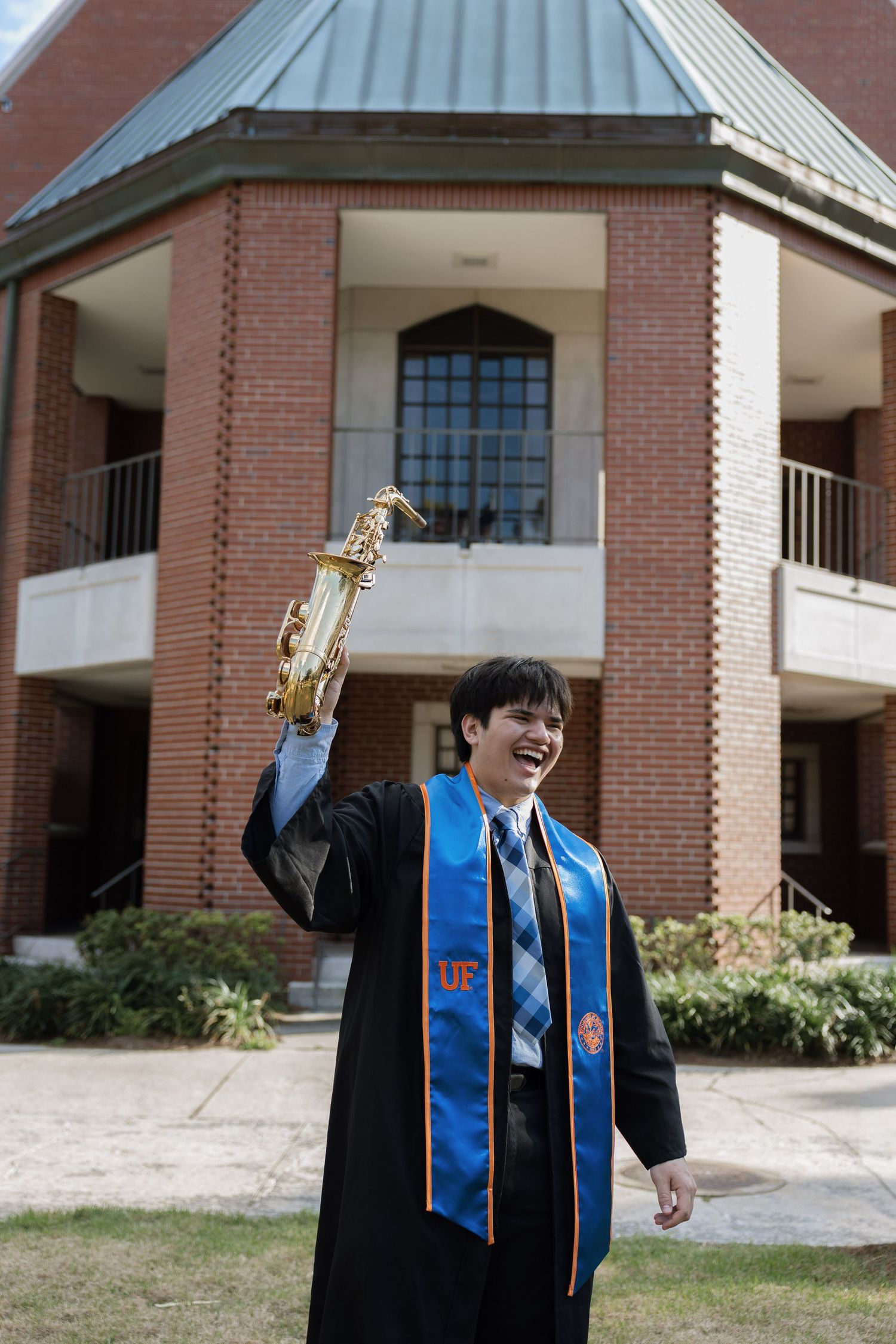UF graduate in cap and gown raises saxophone triumphantly outside brick university building.