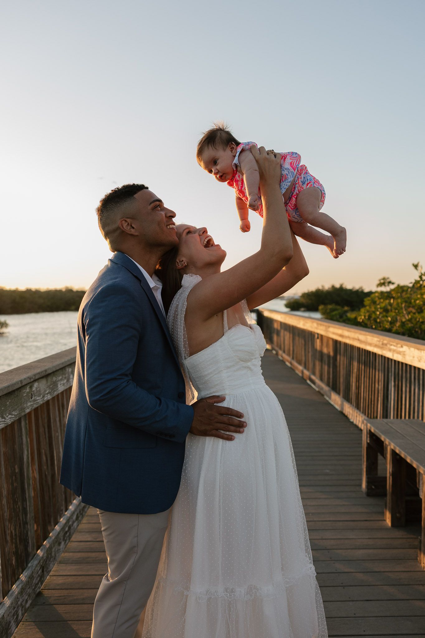 A couple in wedding attire joyfully lifts their baby on a wooden boardwalk at sunset.