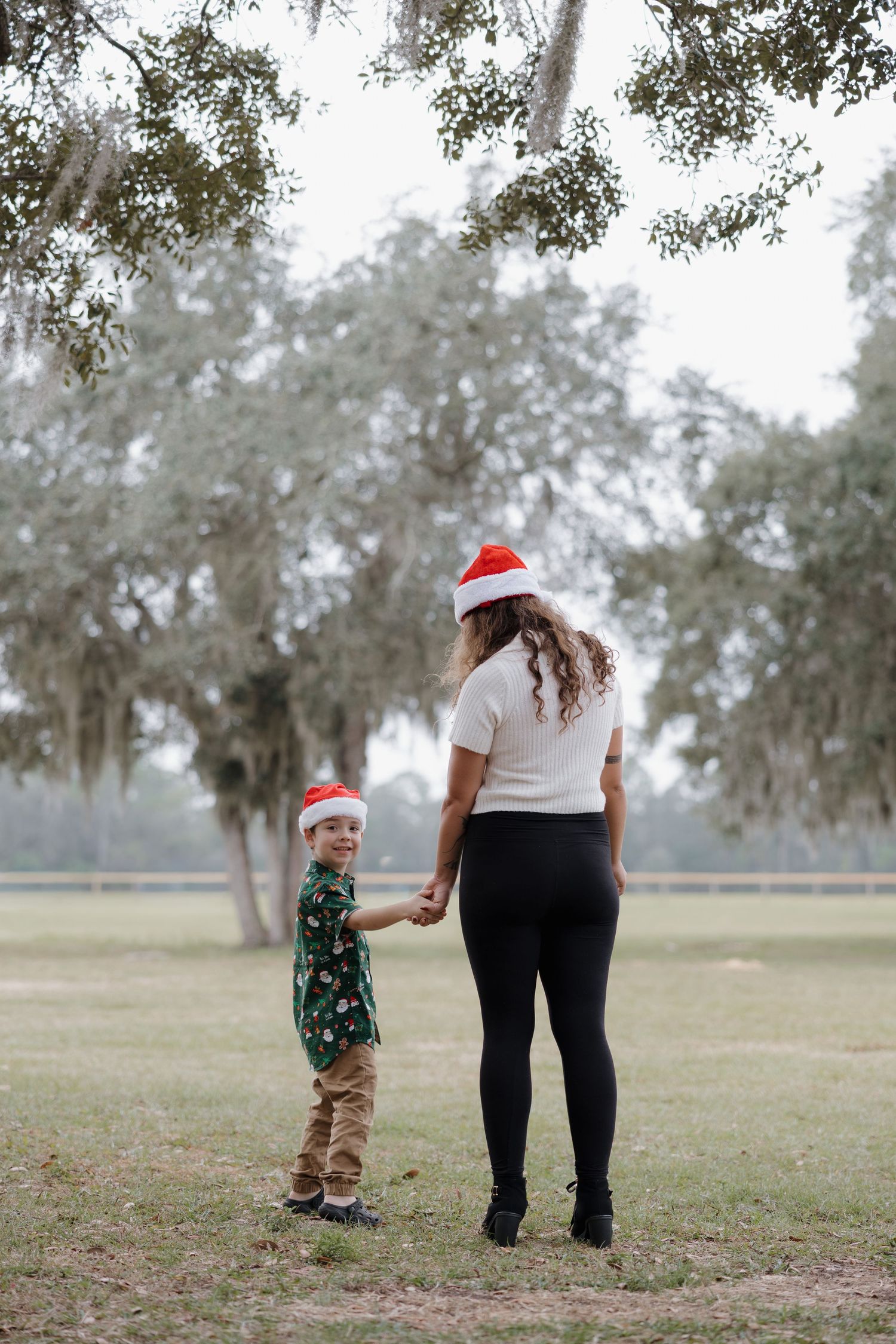 A toddler in a Santa hat walks through a grassy field on a cloudy day.