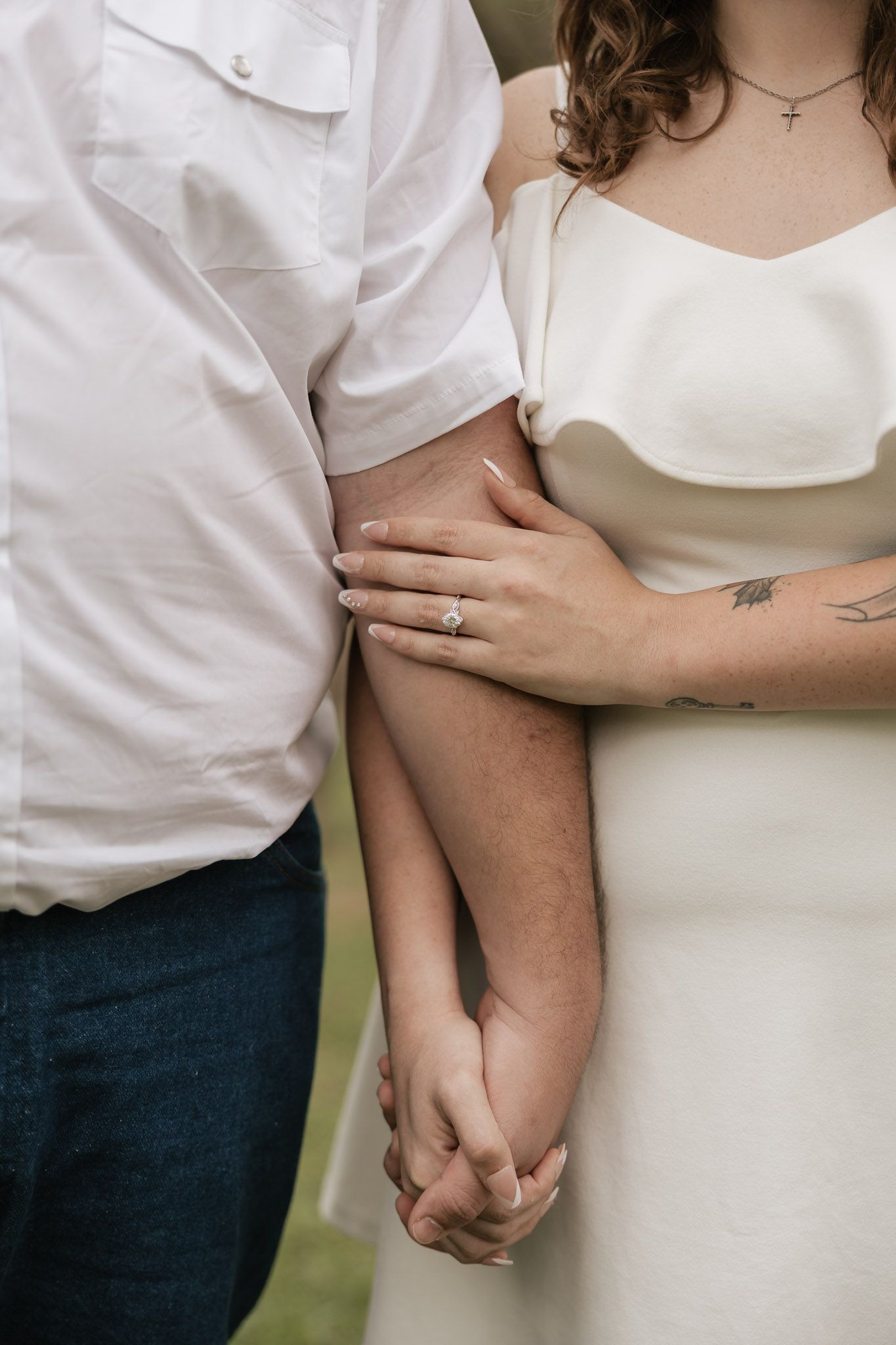 Bride and groom holding hands, showcasing diamond engagement ring, wedding dress, cross necklace, and arm tattoo.