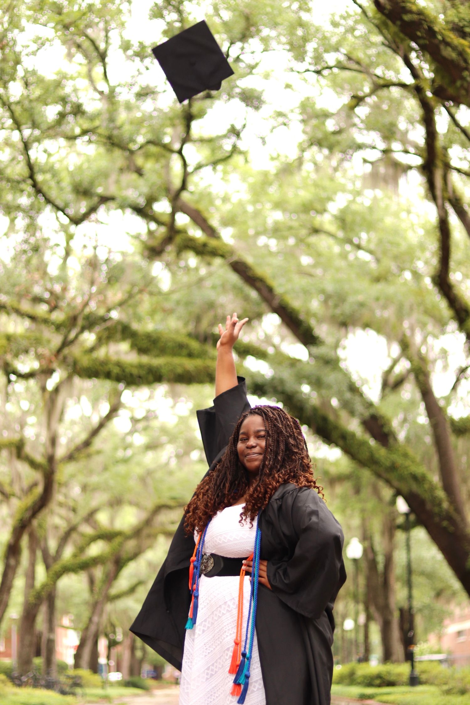 Graduate in black academic regalia celebrates by tossing cap in air among oak trees.