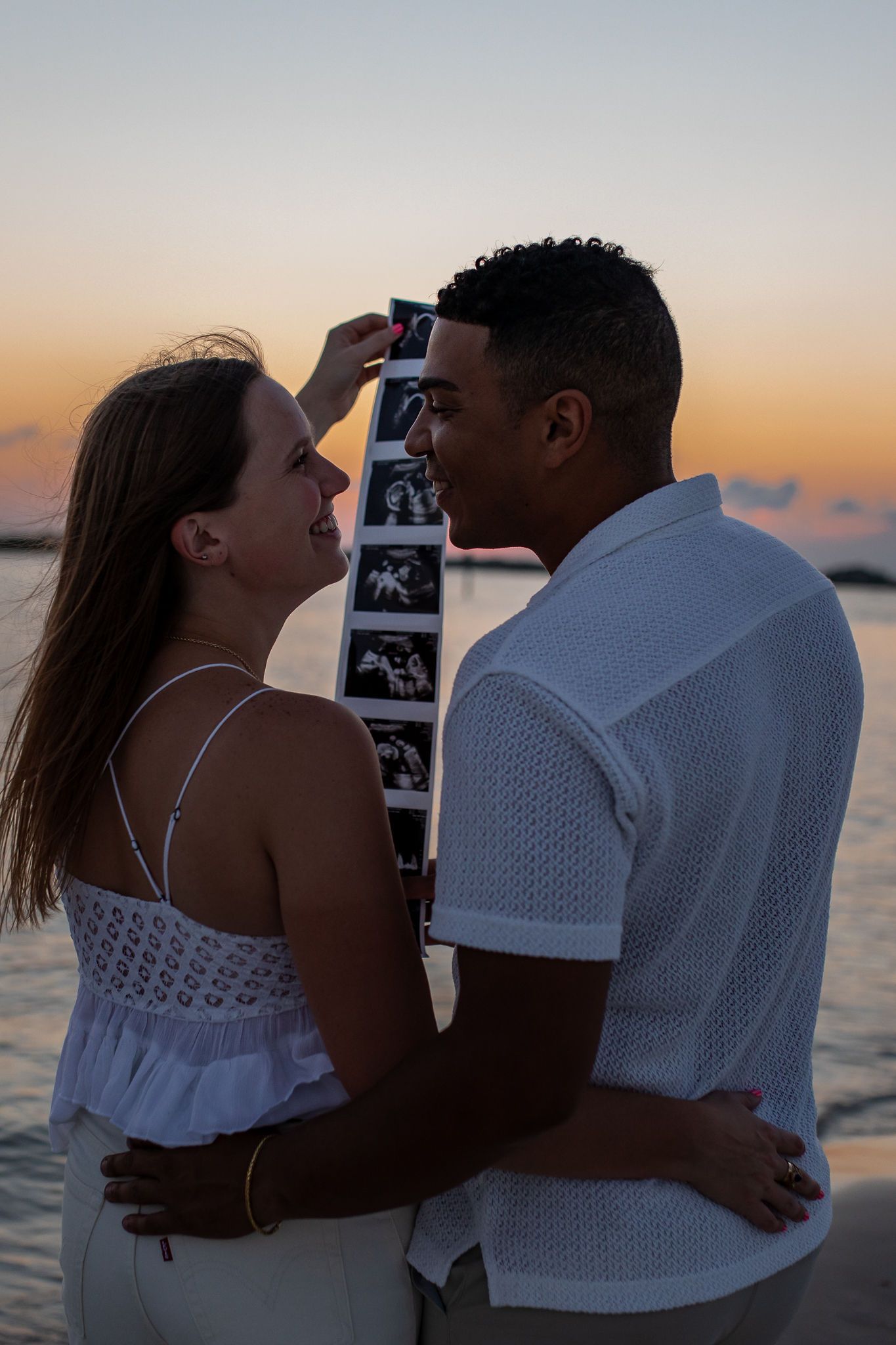 Couple embracing on beach while holding strip of ultrasound photos.