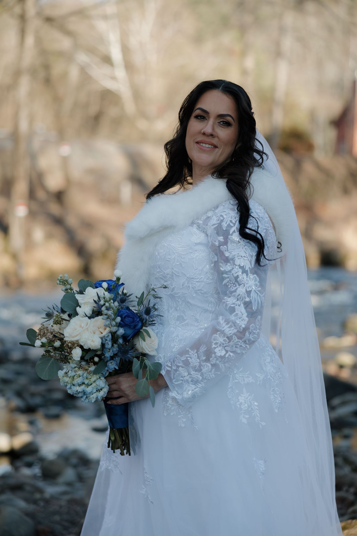 Bride in an elegant white lace wedding gown holding a blue and white bouquet outdoors.