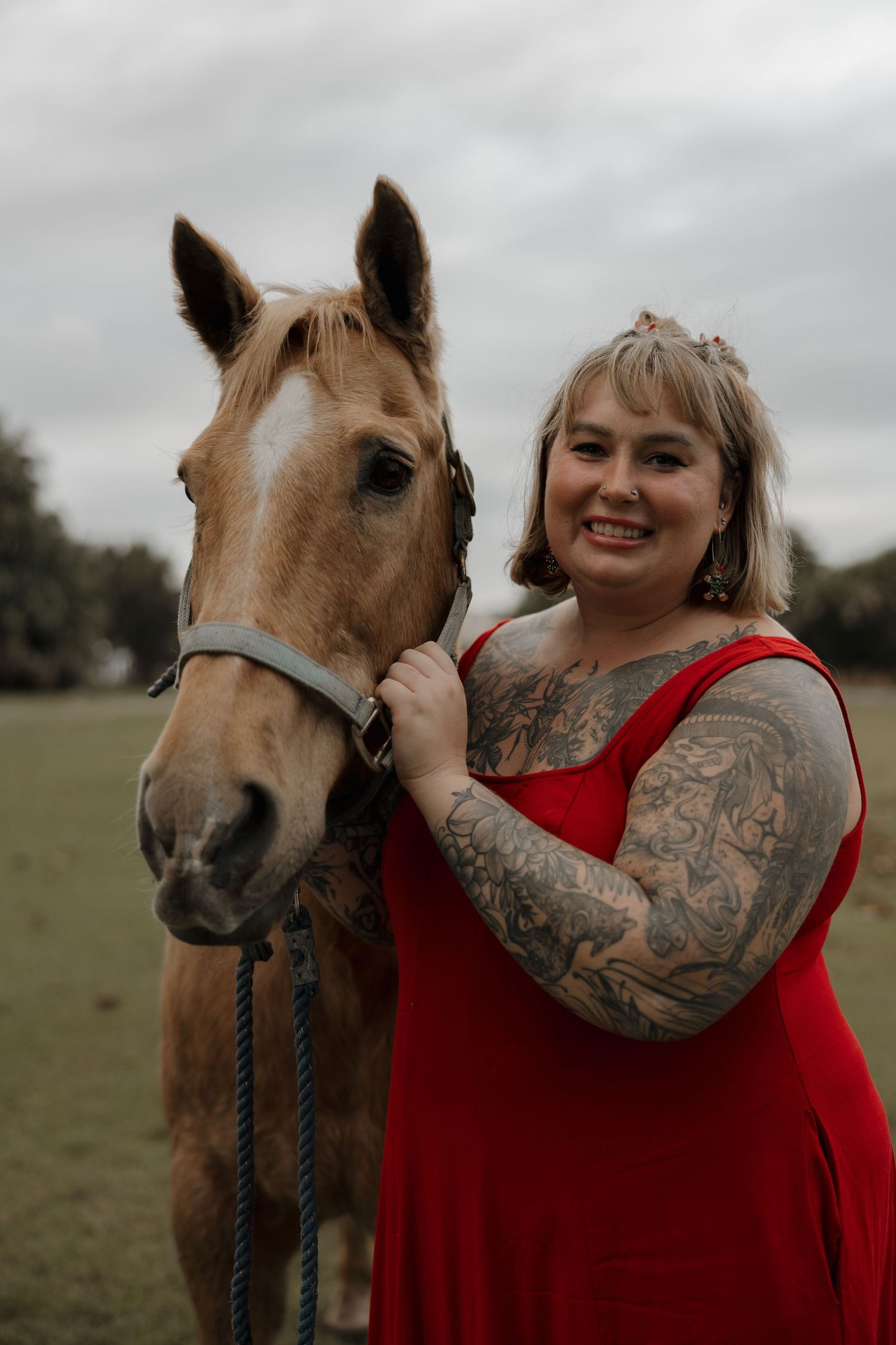 Woman in red top with tattoos poses with a palomino horse in a grassy field.