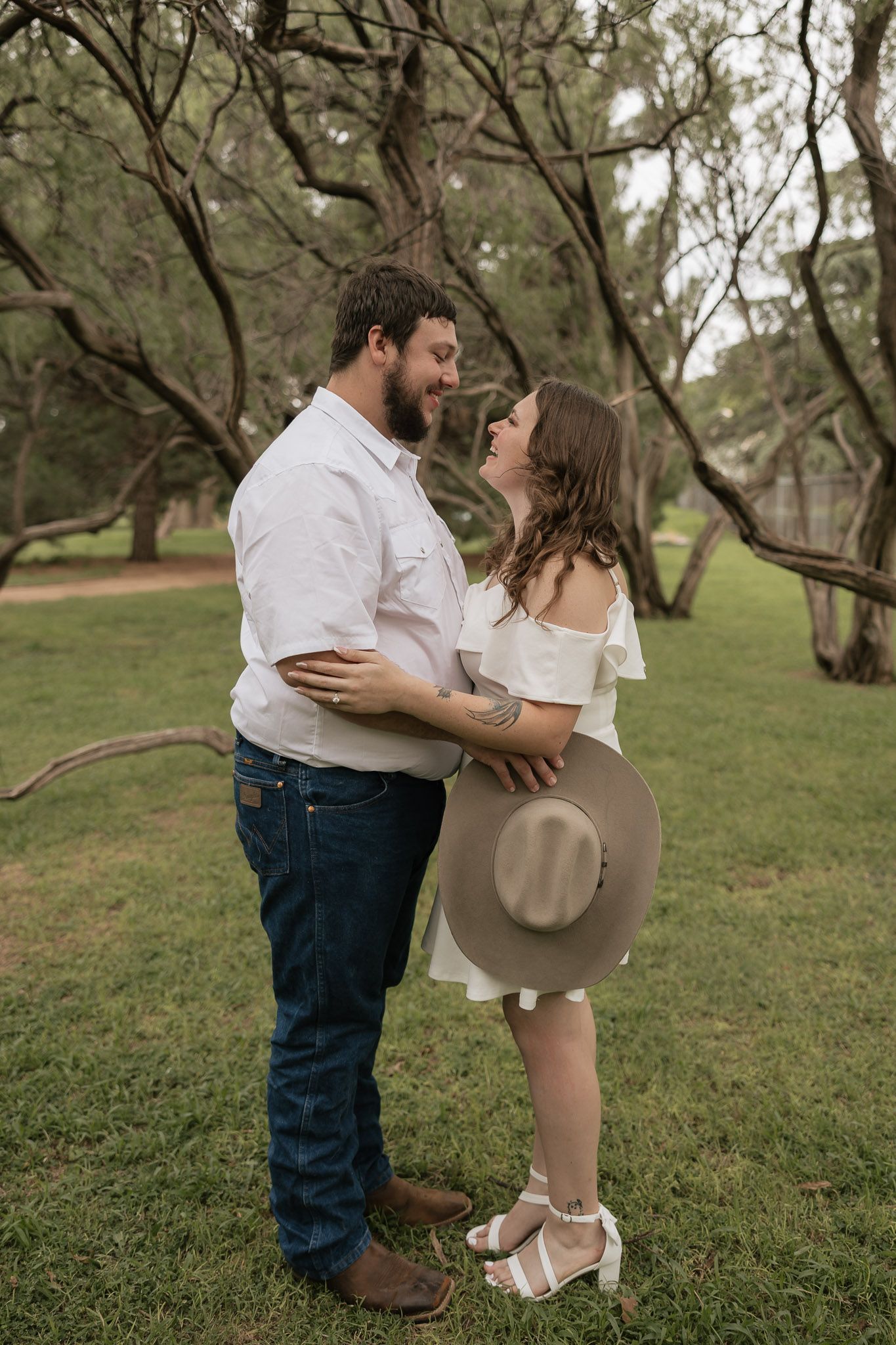 A couple embraces and gazes at each other lovingly in a green park, the woman holding a tan wide-brim hat.