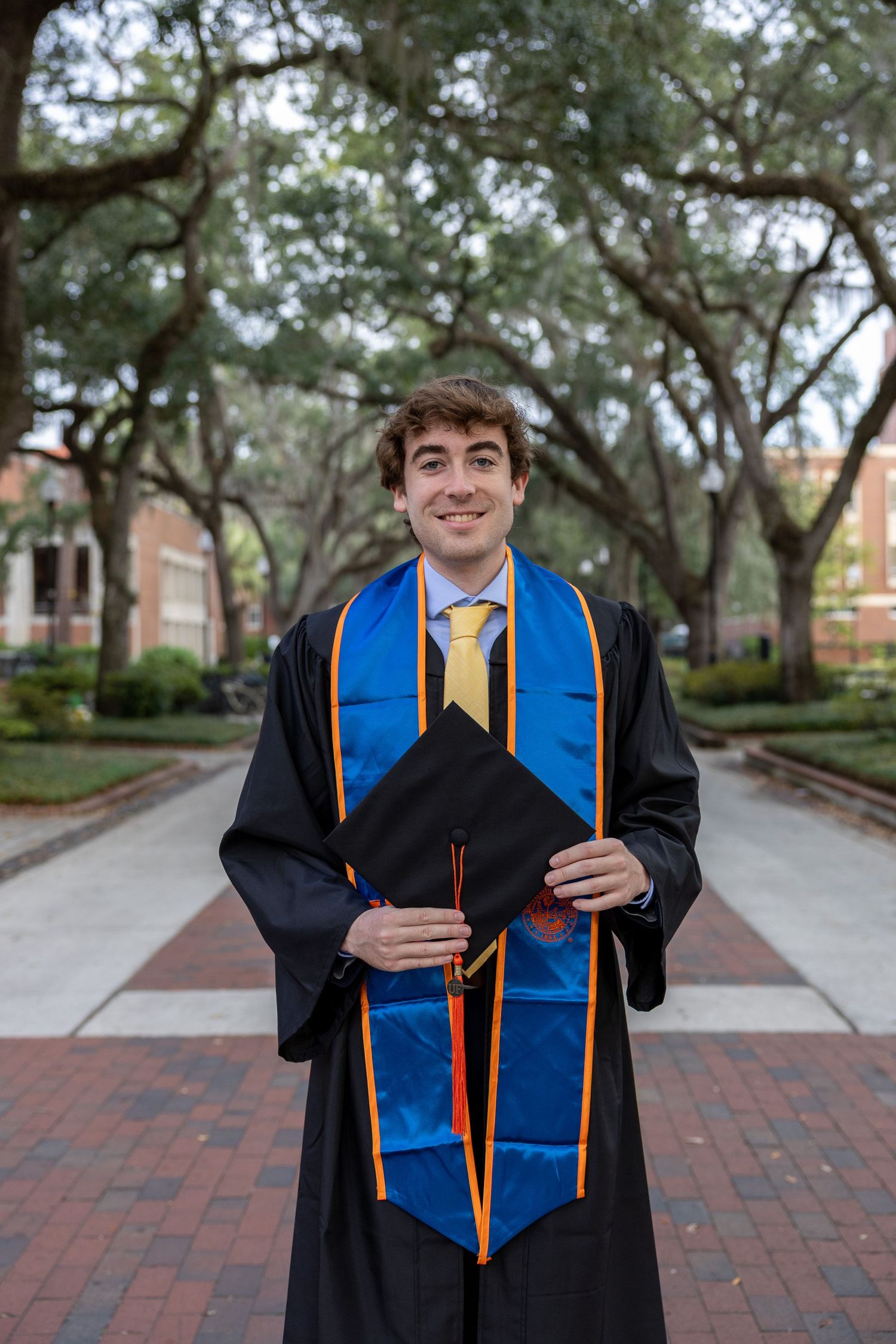Graduate in black cap and gown with blue stole poses on brick pathway.