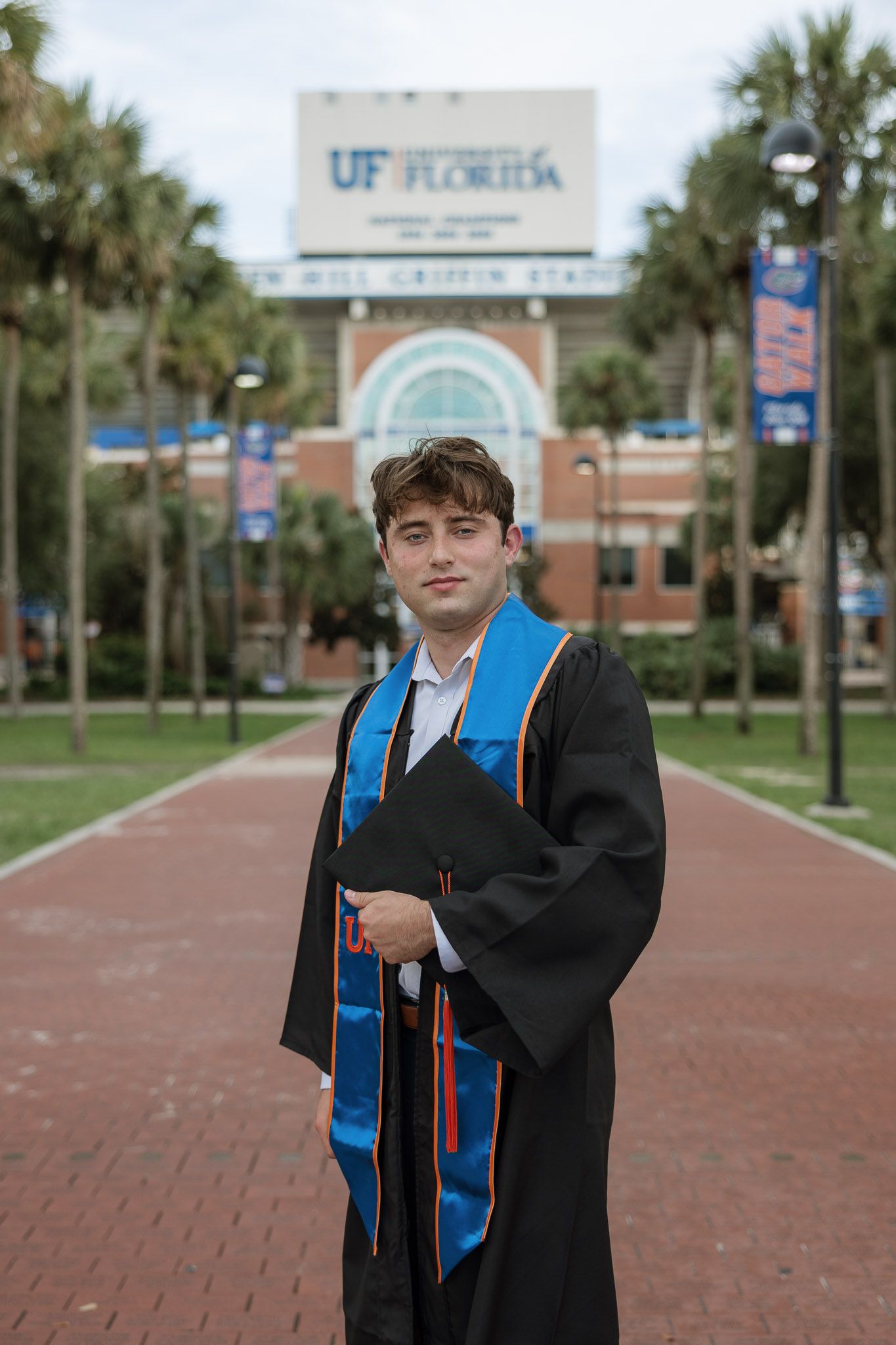 University of Florida graduate in black cap and gown with blue stole poses in front of Ben Hill Griffin Stadium.