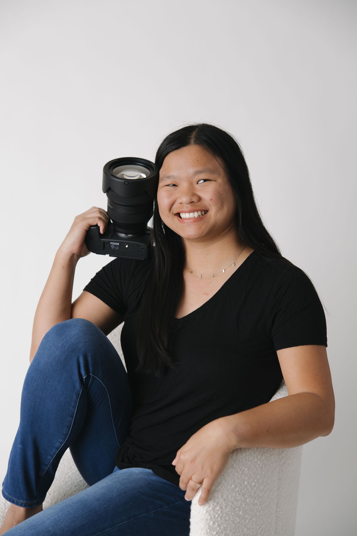 A photographer wearing a black t-shirt and jeans sits casually with their camera against a light background.