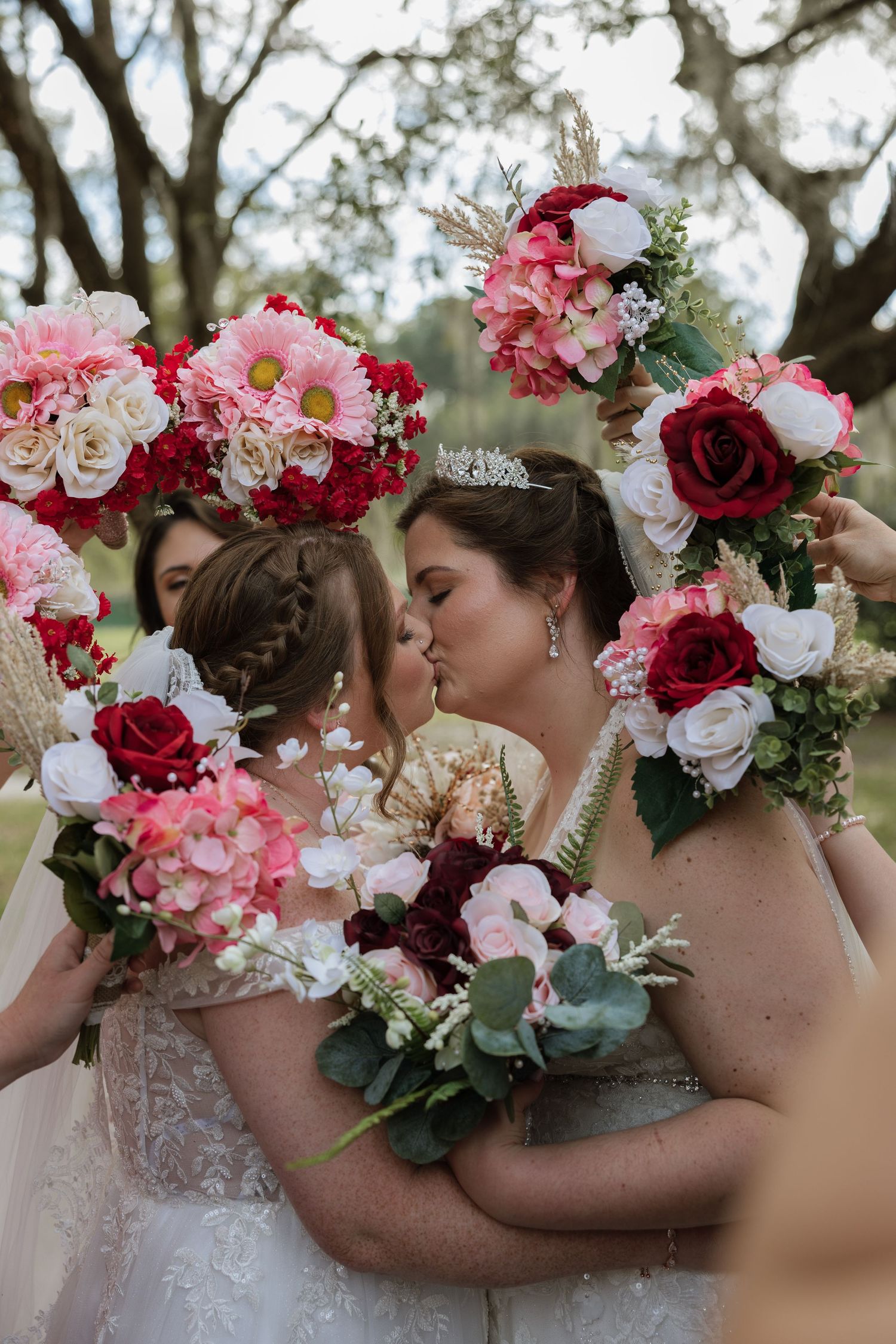 Two brides share a kiss framed by colorful floral bouquets held by bridesmaids at an outdoor wedding ceremony.