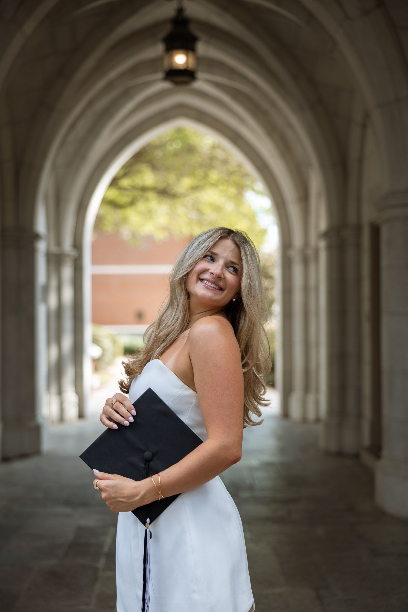 Smiling blonde woman in white dress holding graduation cap under stone archway at university campus.
