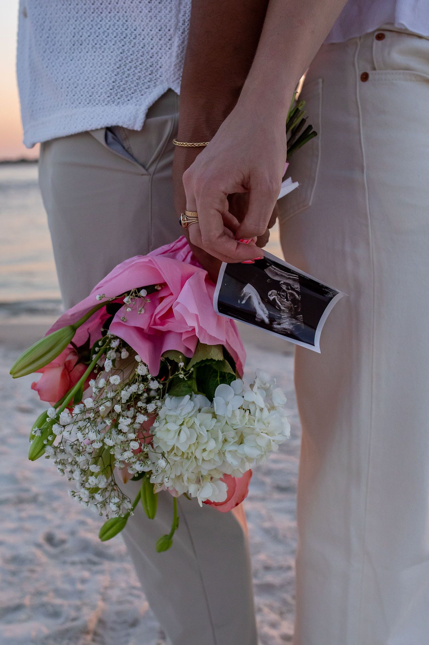 Hands holding pink floral bouquet and ultrasound photo against beach backdrop.