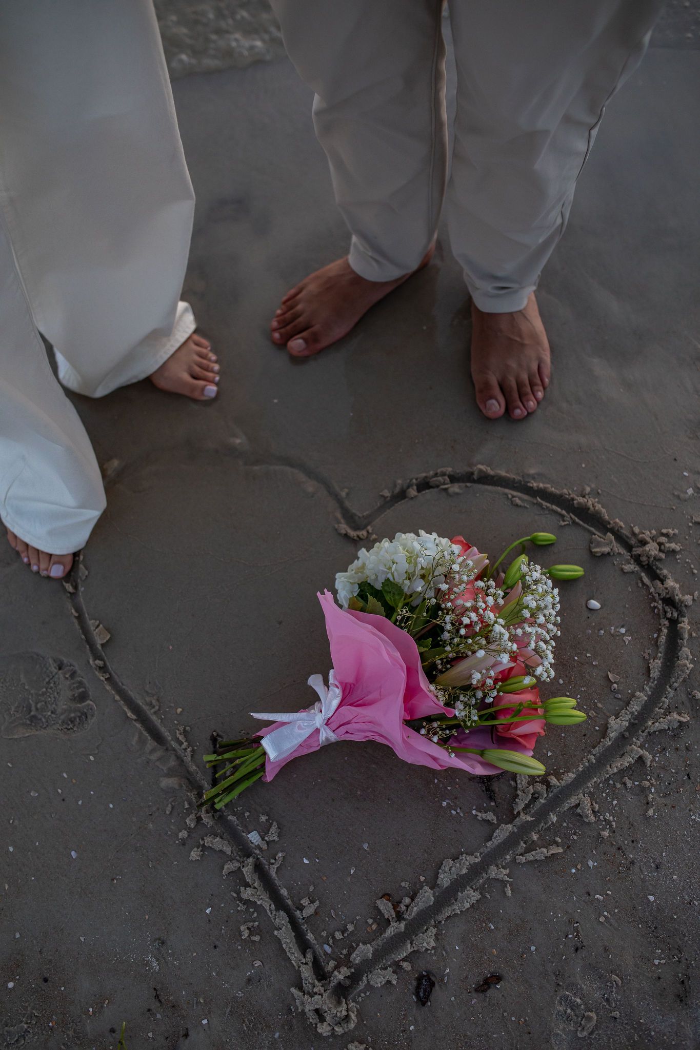 Heart drawn in sand with flowers and bare feet on beach.