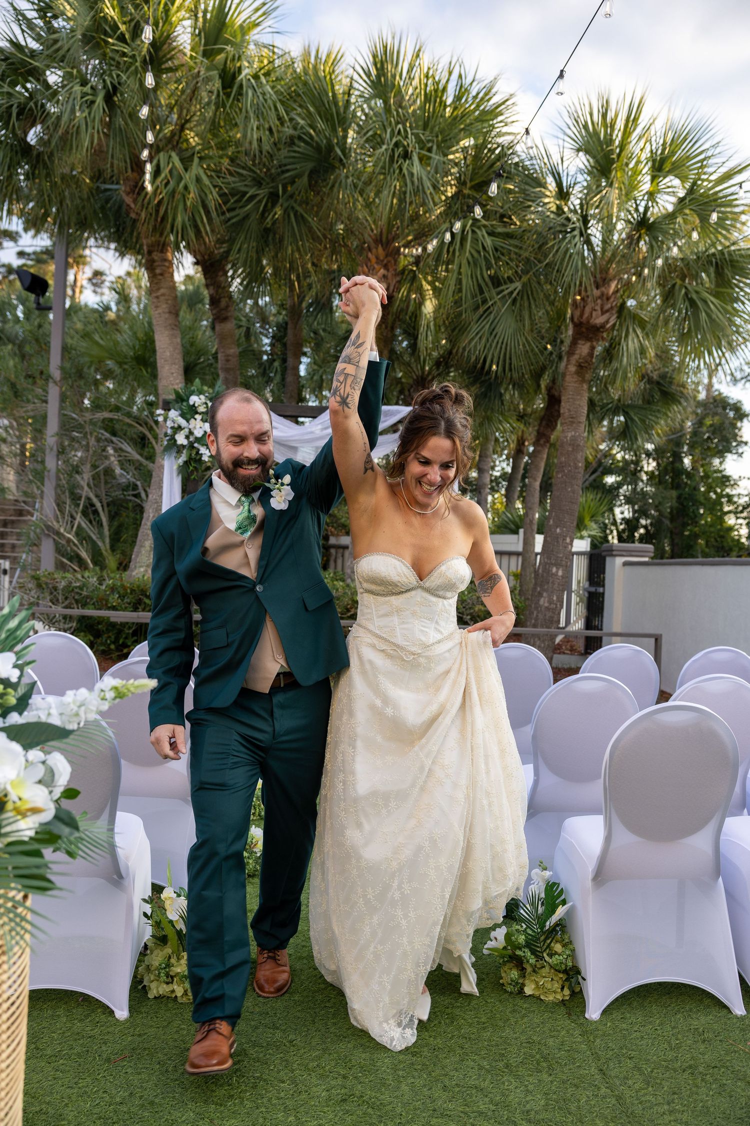 Couple walking together on artificial grass with white-covered chairs and palm trees in background.