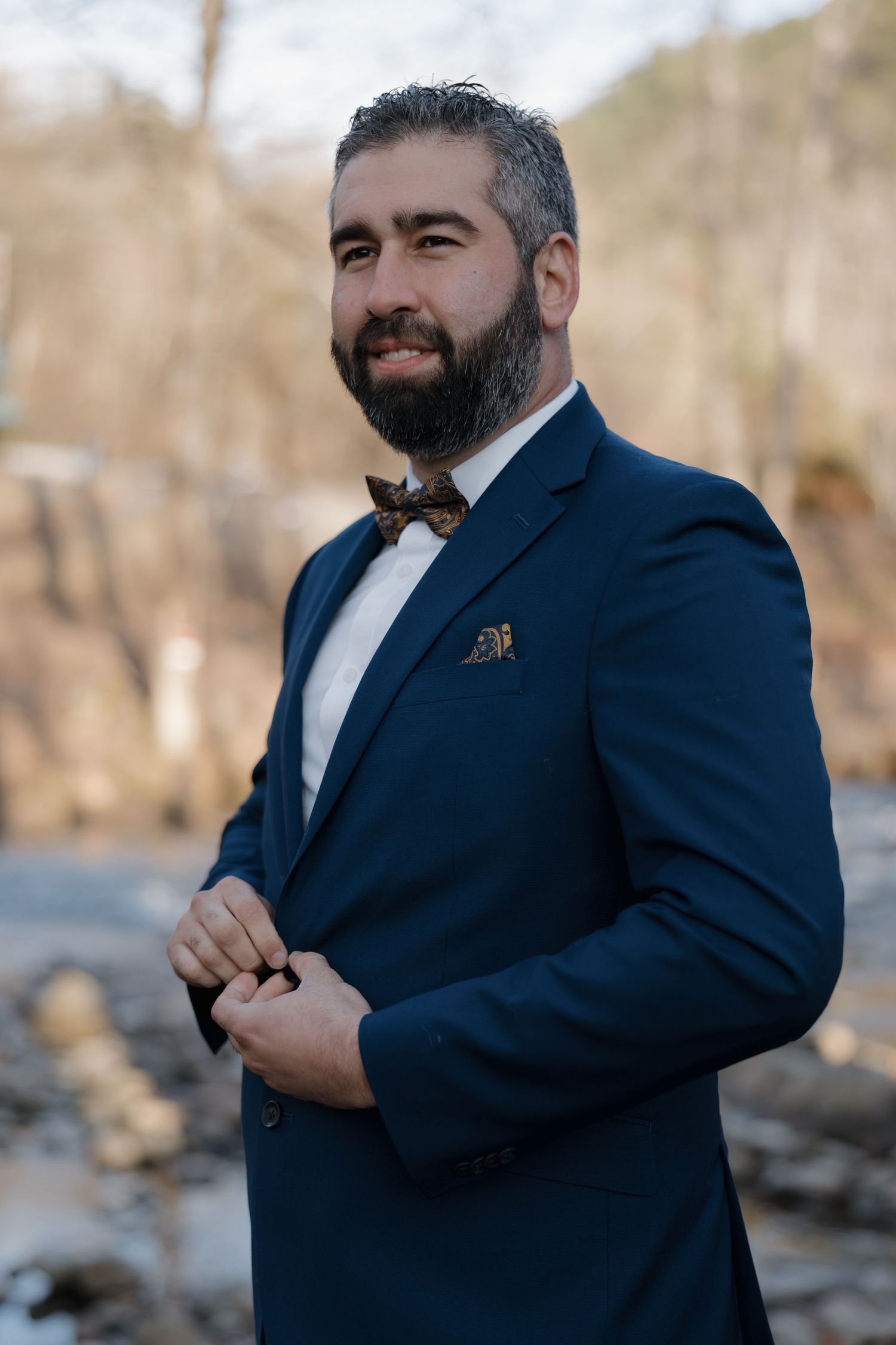 Groom in a navy blue suit poses confidently against a natural outdoor backdrop.