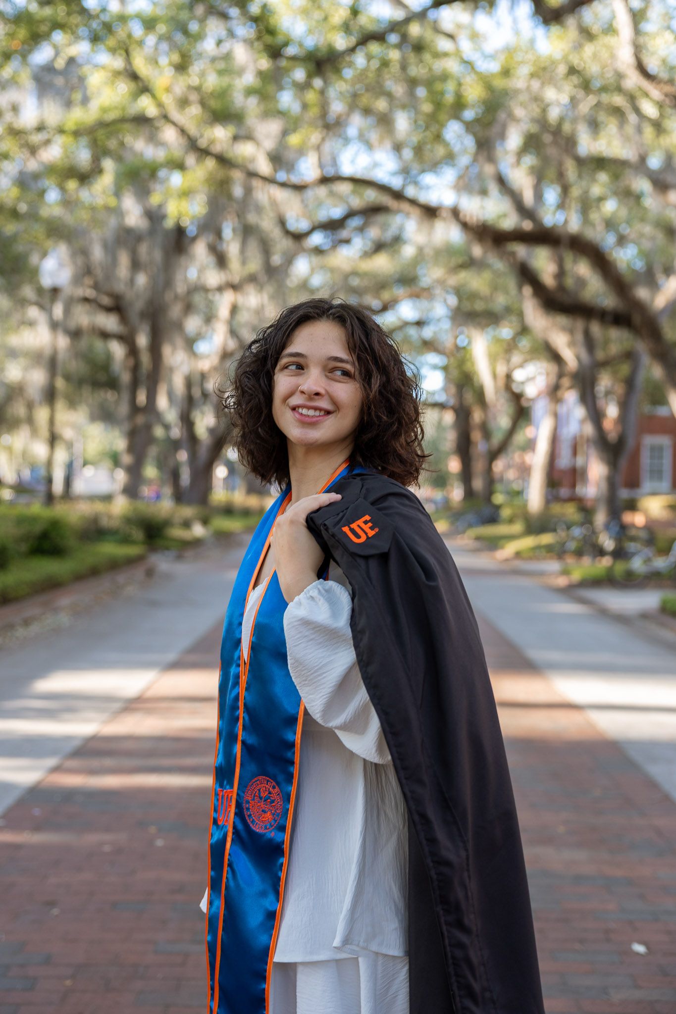 A smiling young woman in a UF graduation gown and blue sash poses on a brick path surrounded by oak trees.