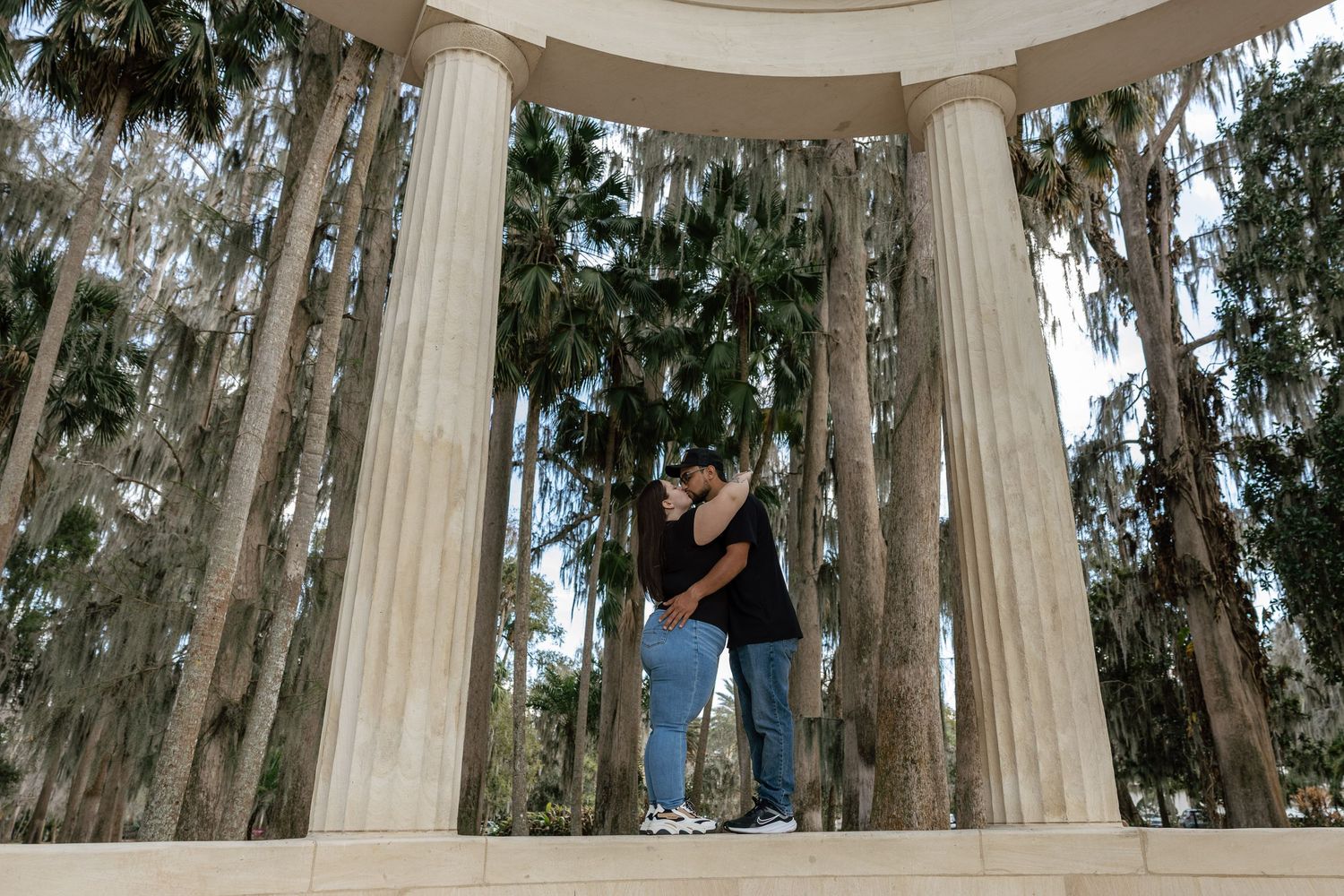 A couple embraces under a white columned gazebo surrounded by Spanish moss-draped trees in a romantic outdoor setting.