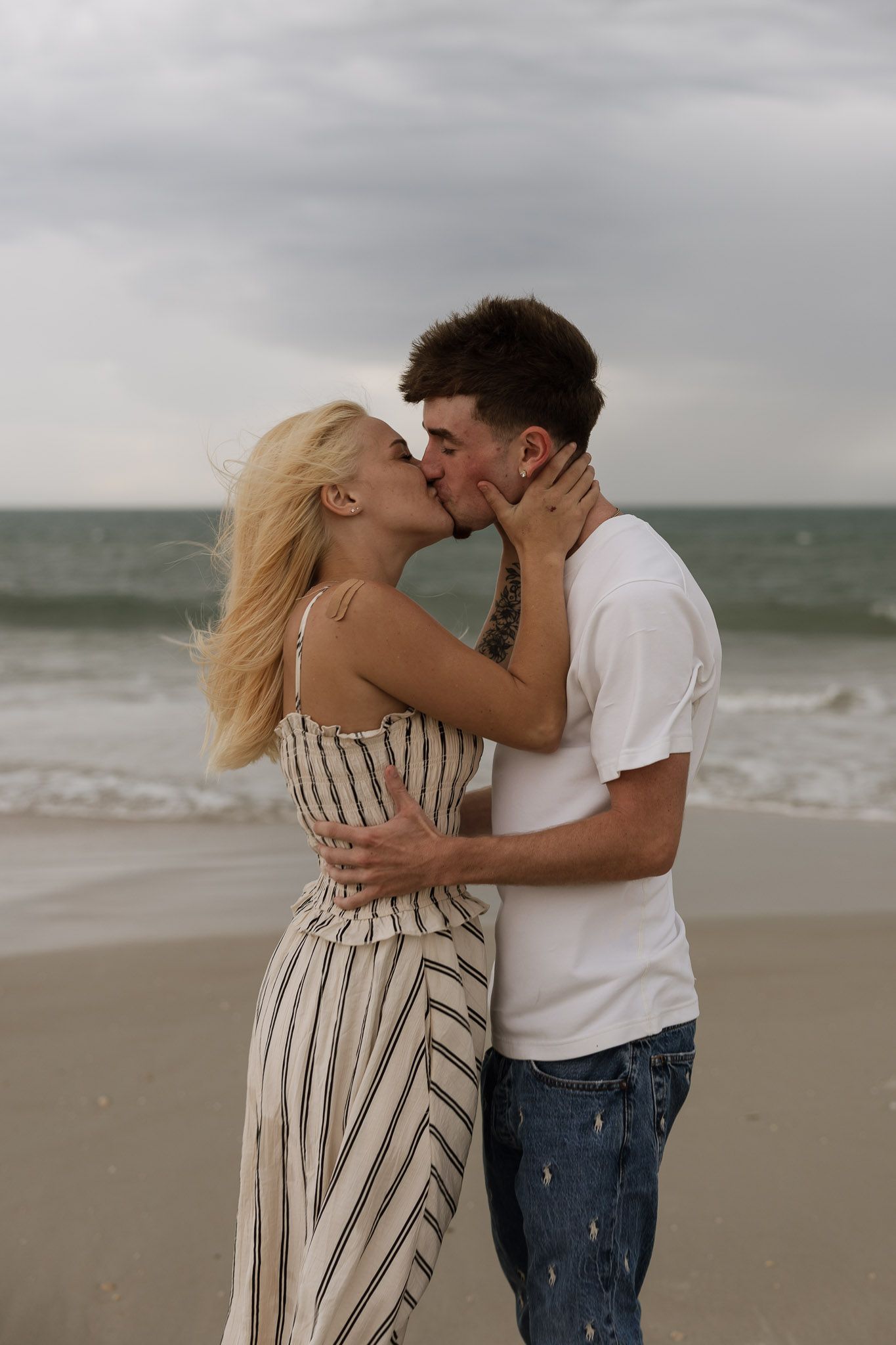 A couple shares a romantic kiss on a beach, with ocean waves in the background on a cloudy day.