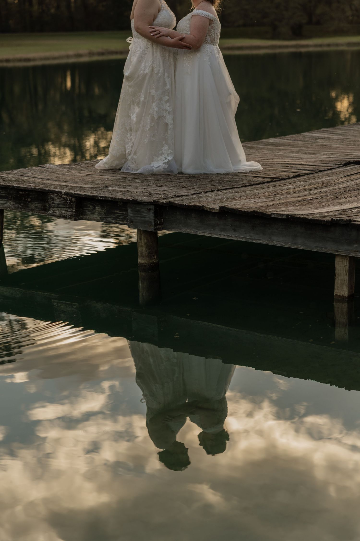 Two brides in white gowns embrace on a wooden dock, their reflection mirrored in the calm lake below.