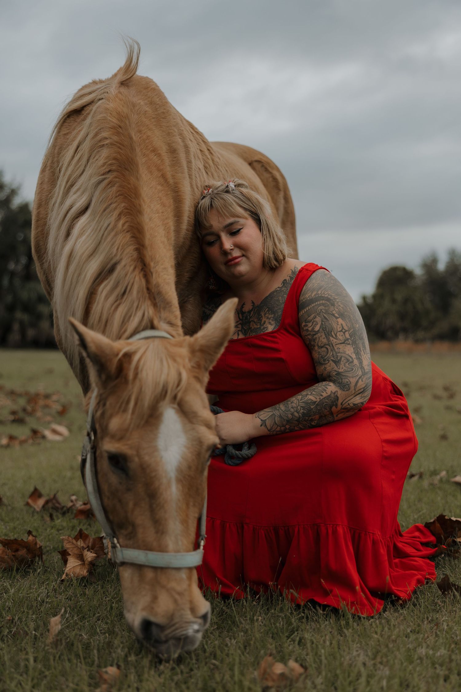A person in a flowing red dress sits in a field with a grazing horse.