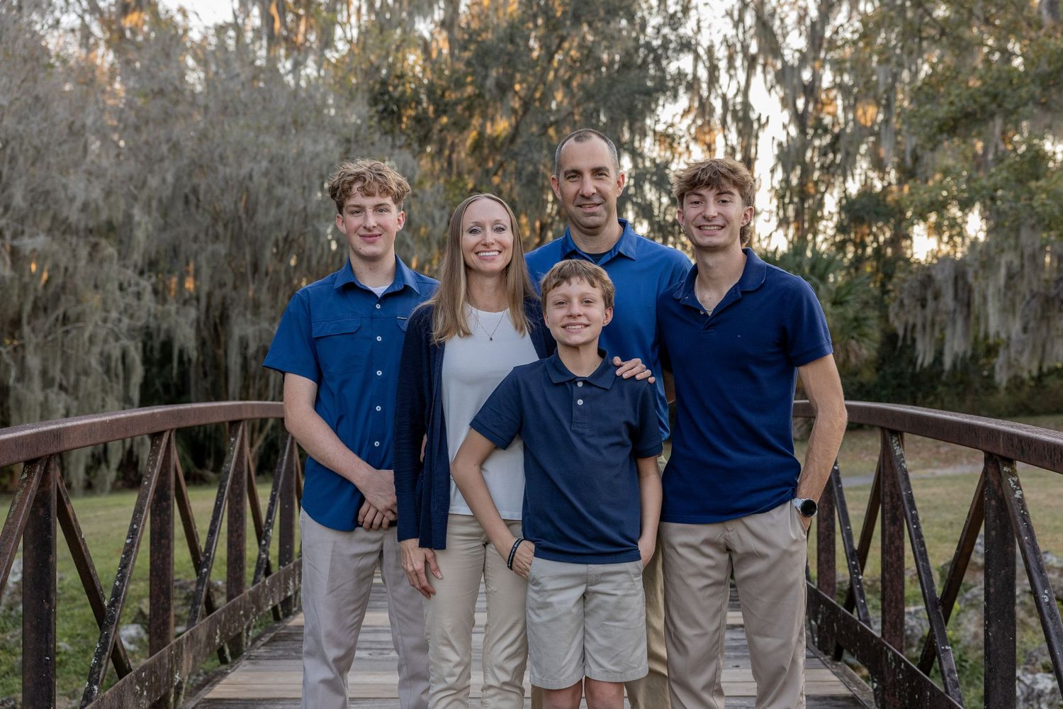 Family wearing coordinated blue and khaki outfits poses together on wooden bridge in nature setting.