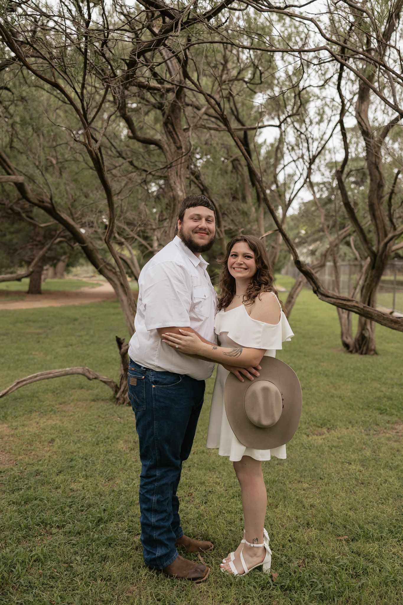 A smiling couple poses outdoors in a park, woman in white dress holding a hat, man in jeans and white shirt.