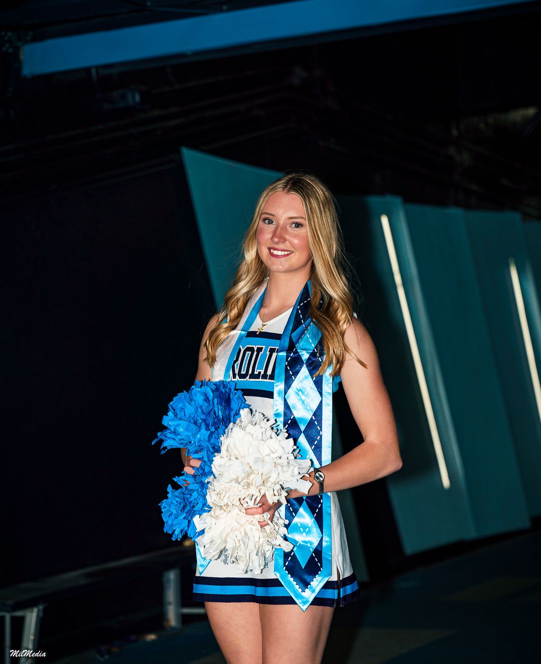 UNC Cheerleader poses with blue and white pom poms in dramatic lighting.