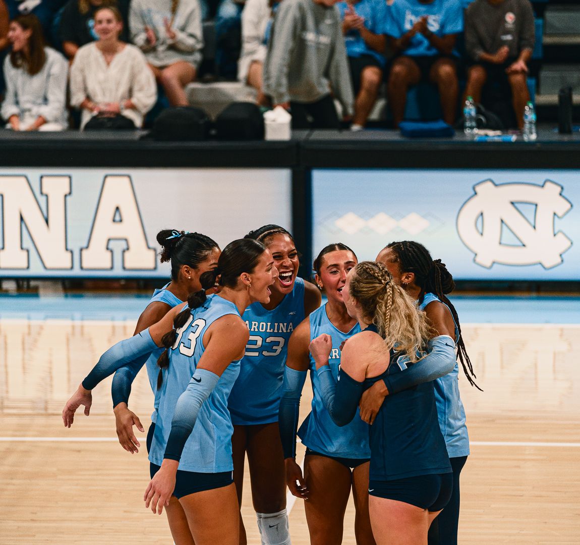 Women's volleyball team members huddle together on court wearing Carolina blue jerseys.