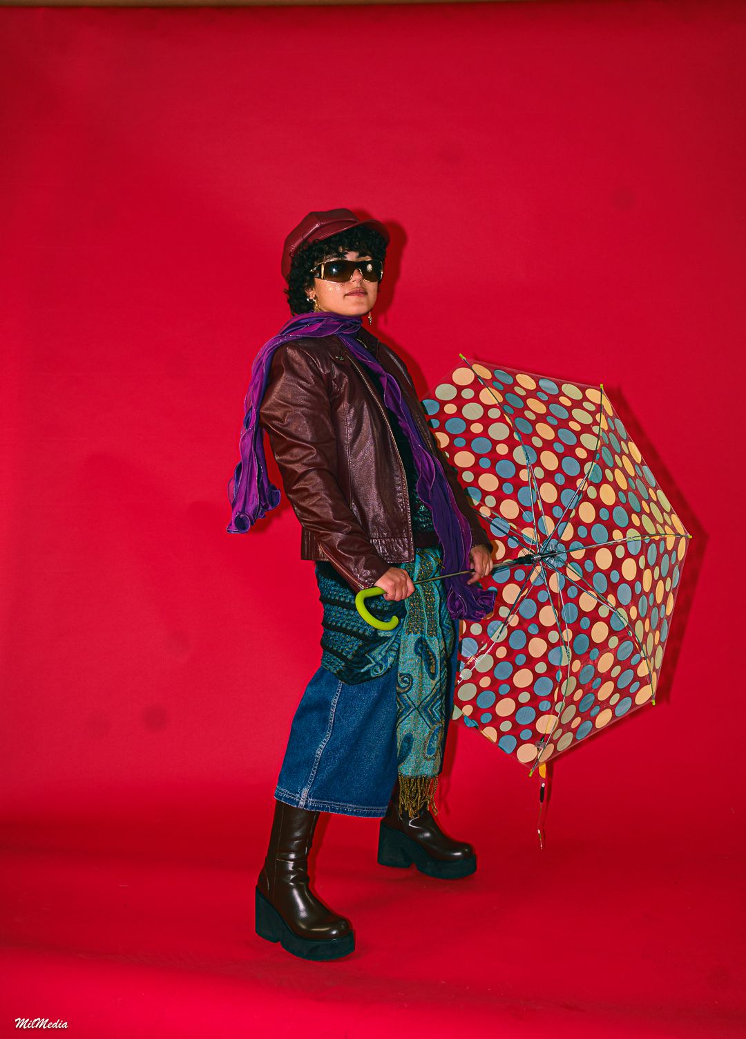 Figure in dark clothing holds colorful polka dot umbrella against red studio backdrop.
