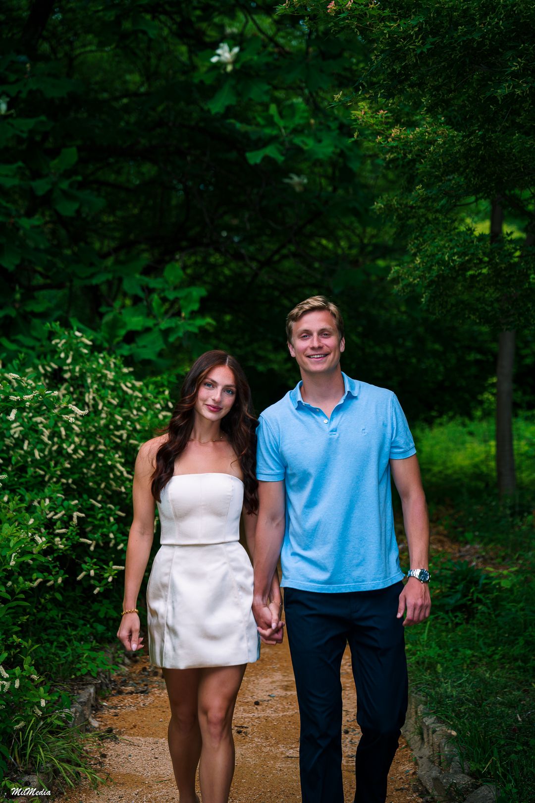 Couple walks together on a wooded trail surrounded by lush green foliage during a graduation shoot in the Coker Arboretum.