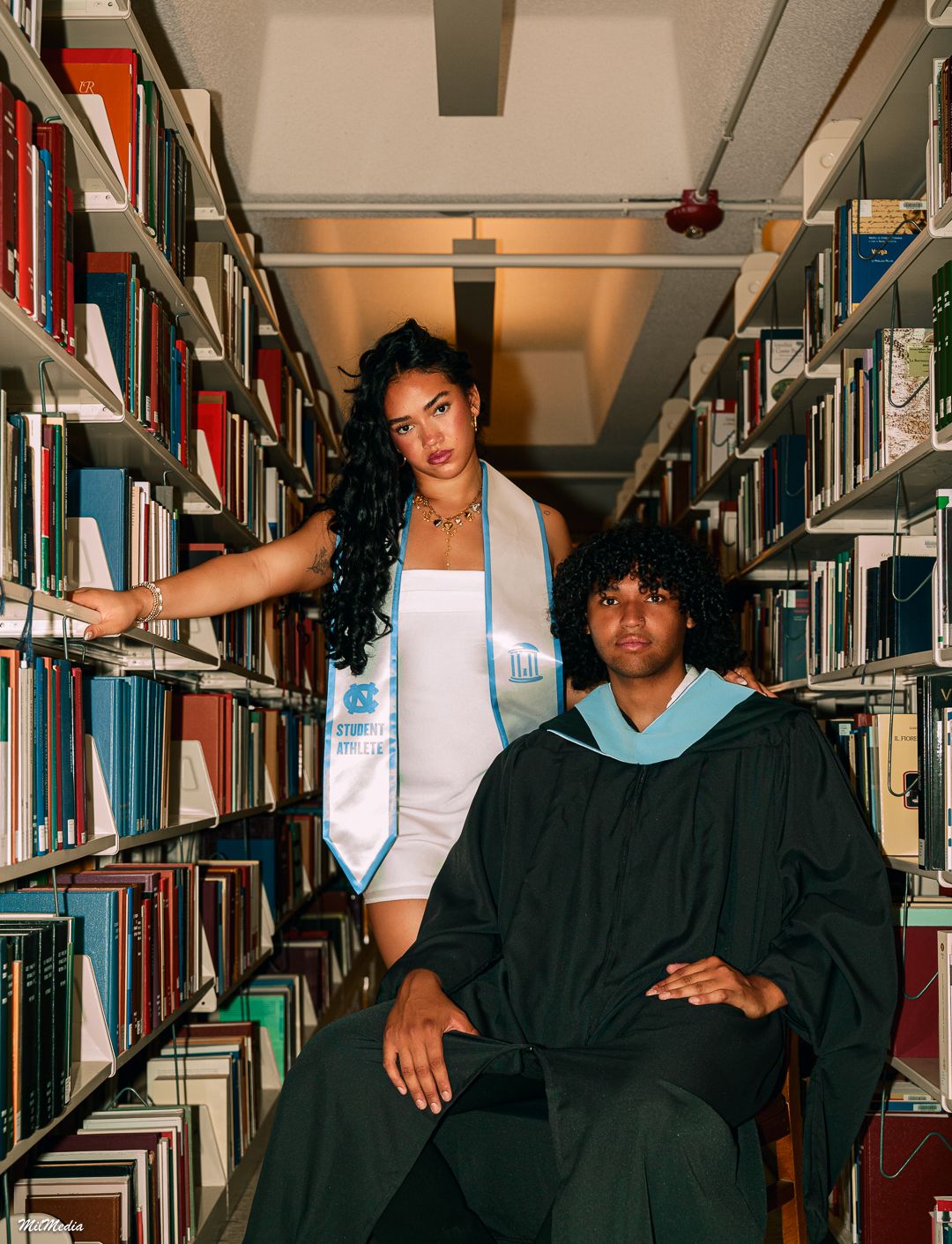 Graduates pose between library bookshelves wearing academic regalia and white dress.
