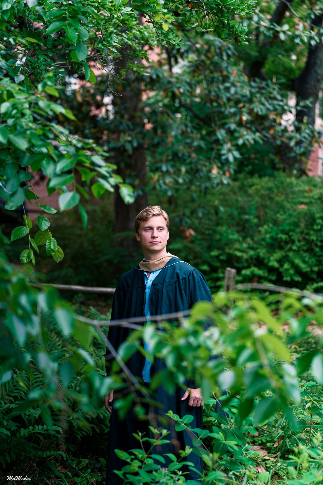 Graduate poses among leafy green foliage wearing traditional black graduation gown in the UNC Coker Arboretum.