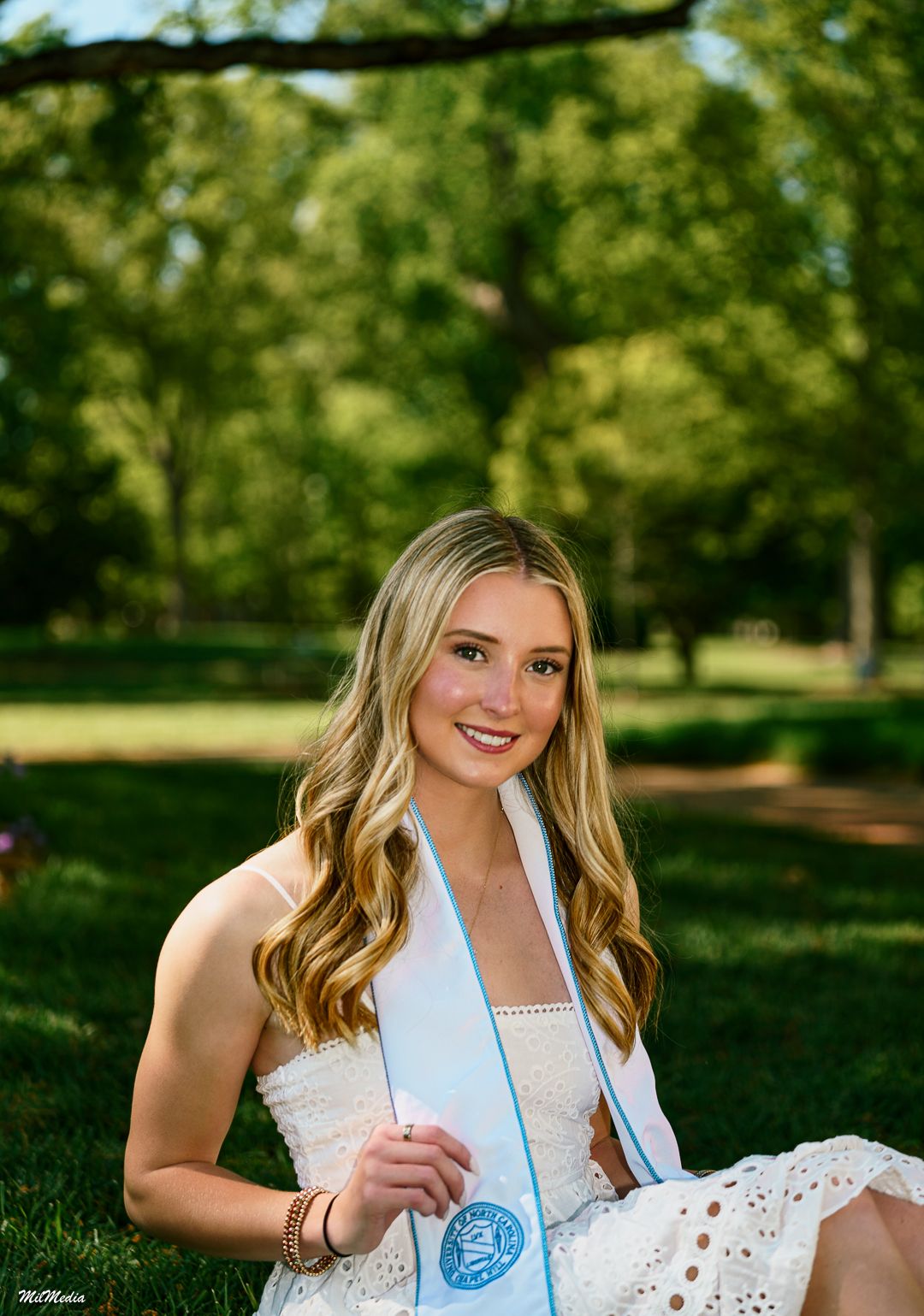 A person in a white eyelet dress poses outdoors in a sunny park setting with green trees in the background.