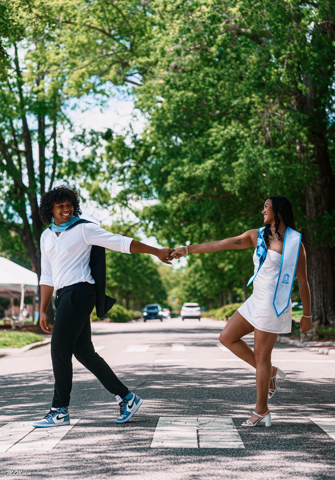 Graduates in blue sashes strike playful pose on tree-lined campus walkway.