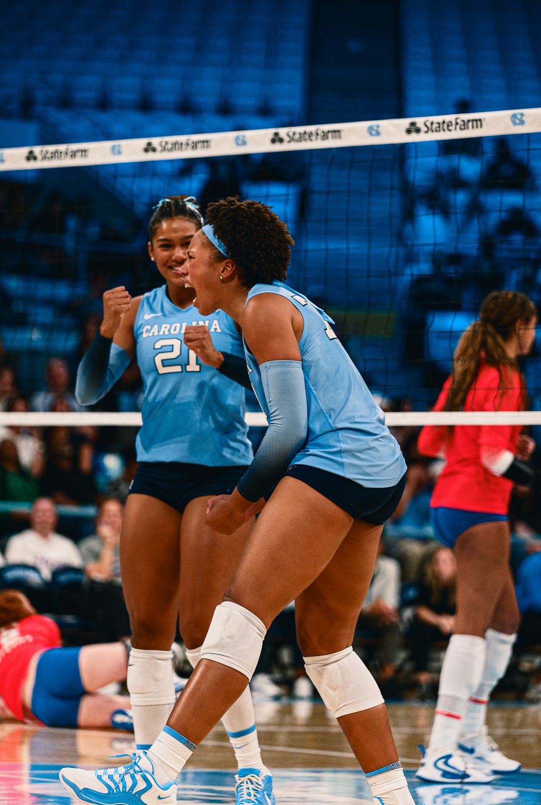 Volleyball players in light blue uniforms crouch in defensive positions during game play.