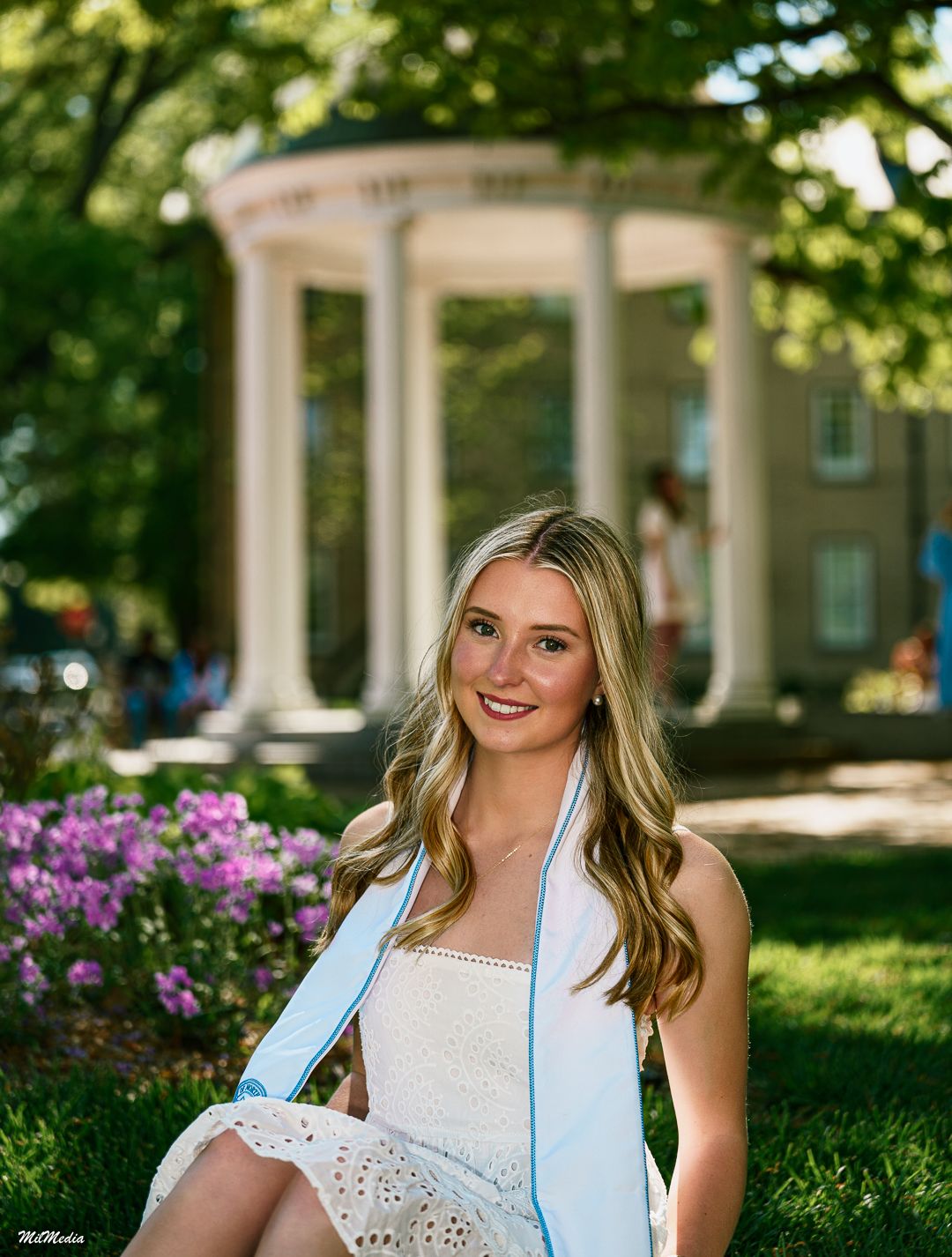 A graduate in white dress poses outdoors with purple flowers and classical architecture.