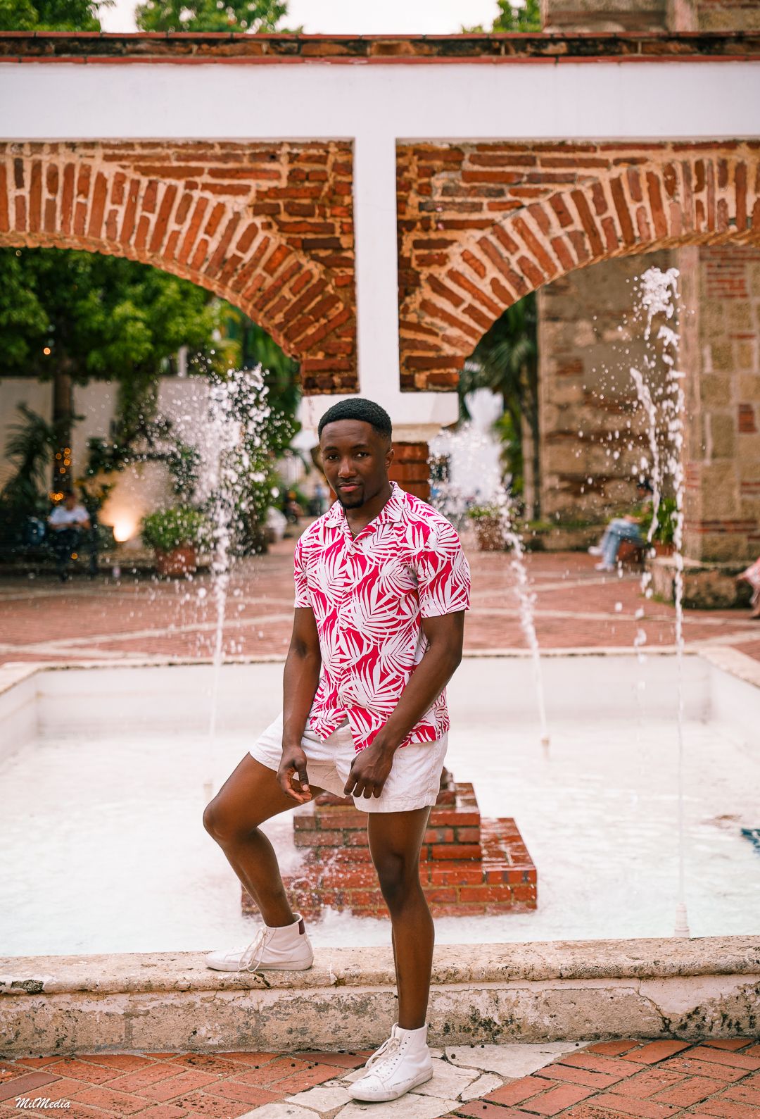 Person in pink patterned shirt poses by fountain with brick arches in background.