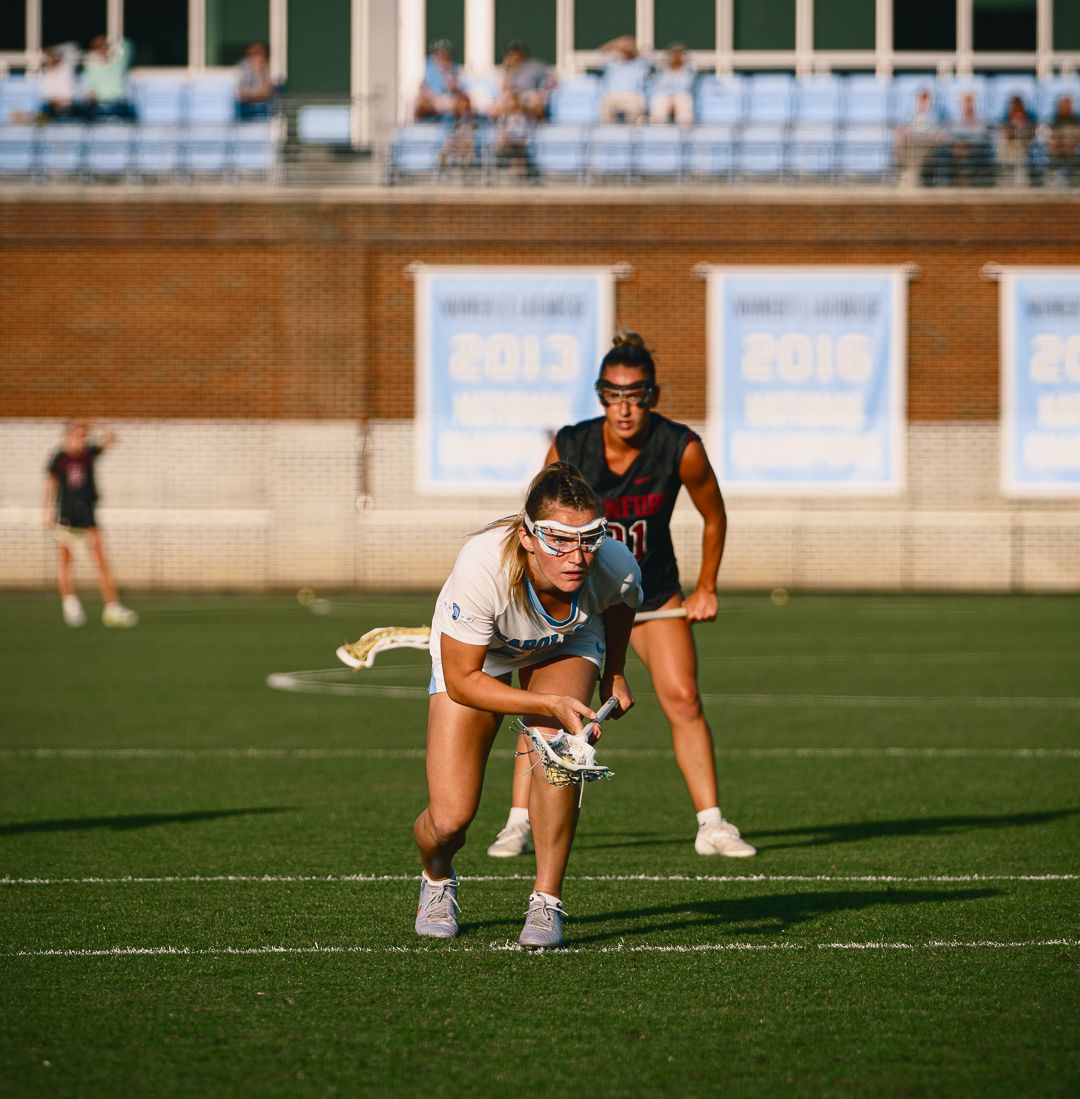 Two female lacrosse players compete on a turf field during a sunny game, one defending the other in white gear.