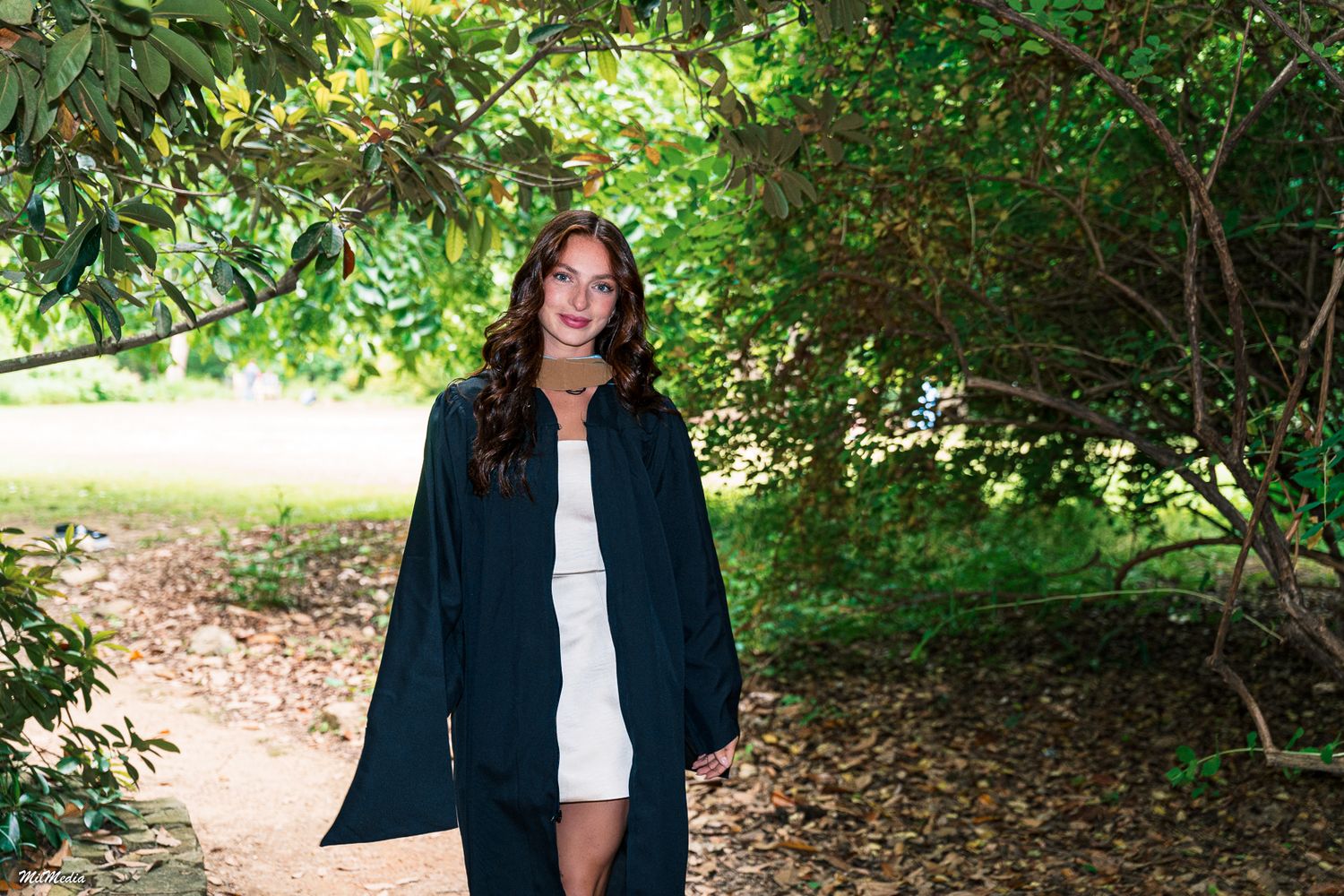 Figure in dark coat and white dress stands on a leaf-covered woodland path.