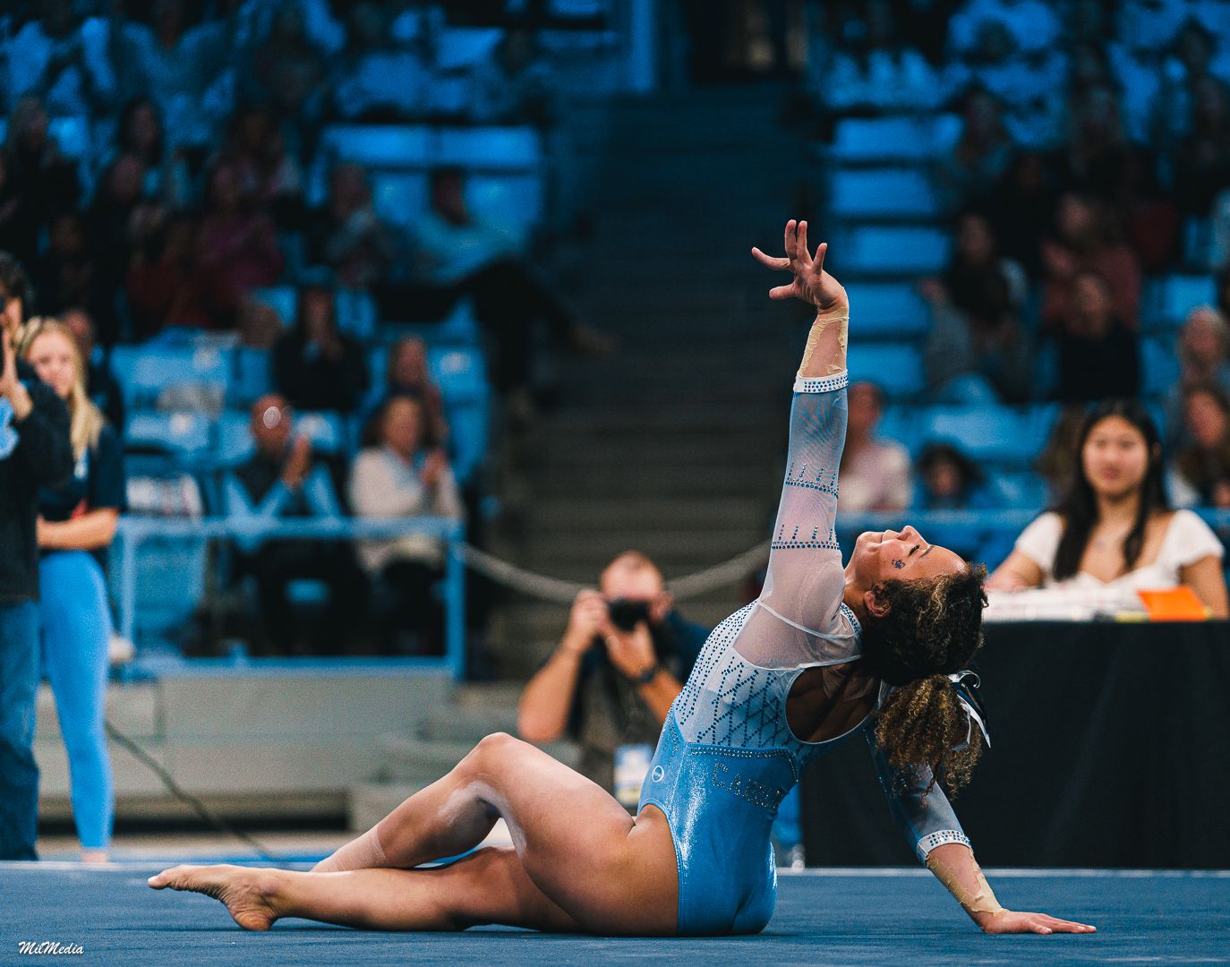 A gymnast performs a graceful floor routine in a light blue leotard at a competitive event with spectators in the background.