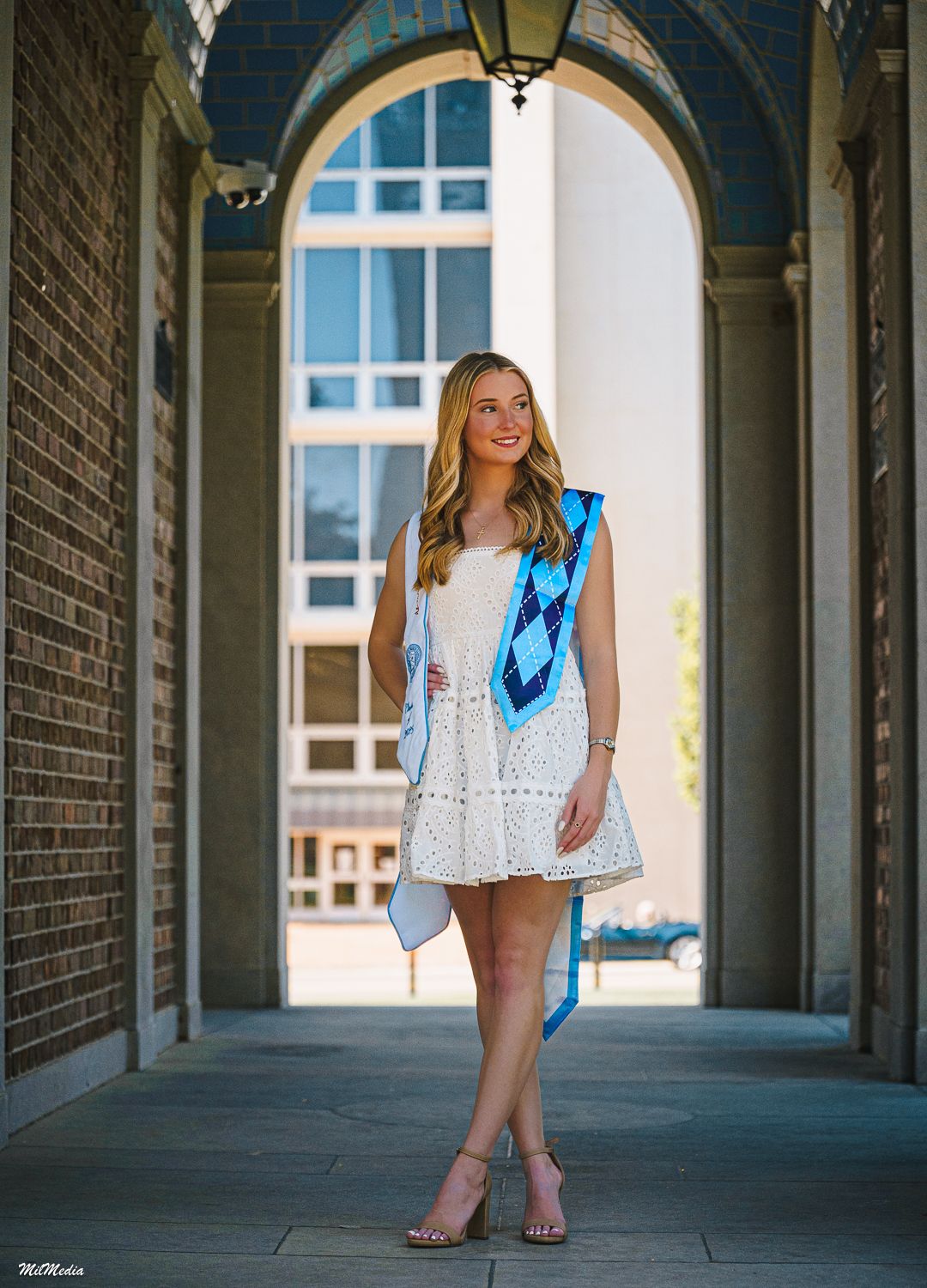 A graduate in a white dress poses in an arched brick colonnade underneath the South Building at UNC