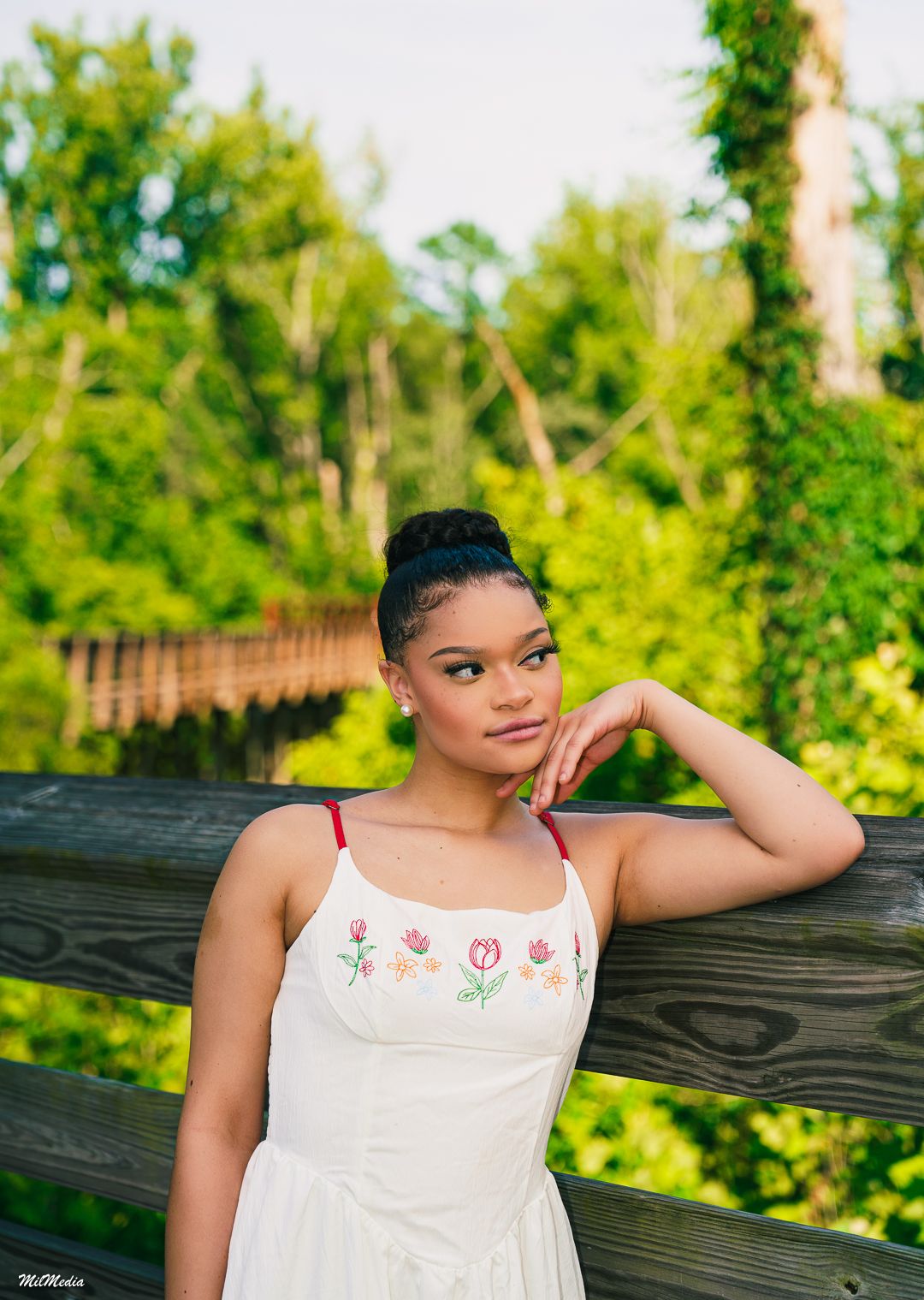 A person in a white embroidered top poses outdoors against a wooden fence with lush greenery in the background.